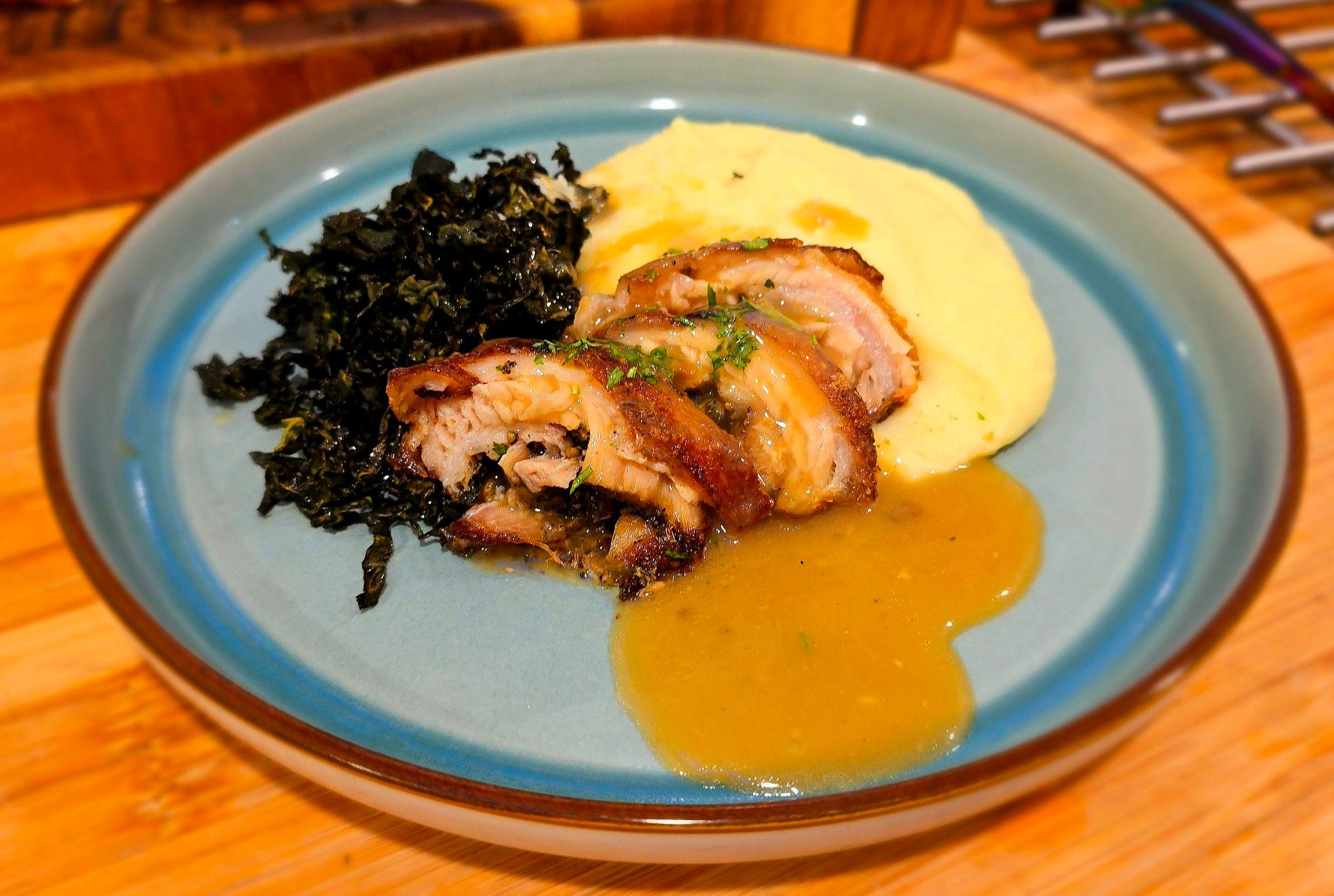 A blue plate, with a pile of golden mashed potato in the center, to the left crispy green cavolo nero, and placed in front three thin slices of pork belly porchetta, with a pale golden gravy