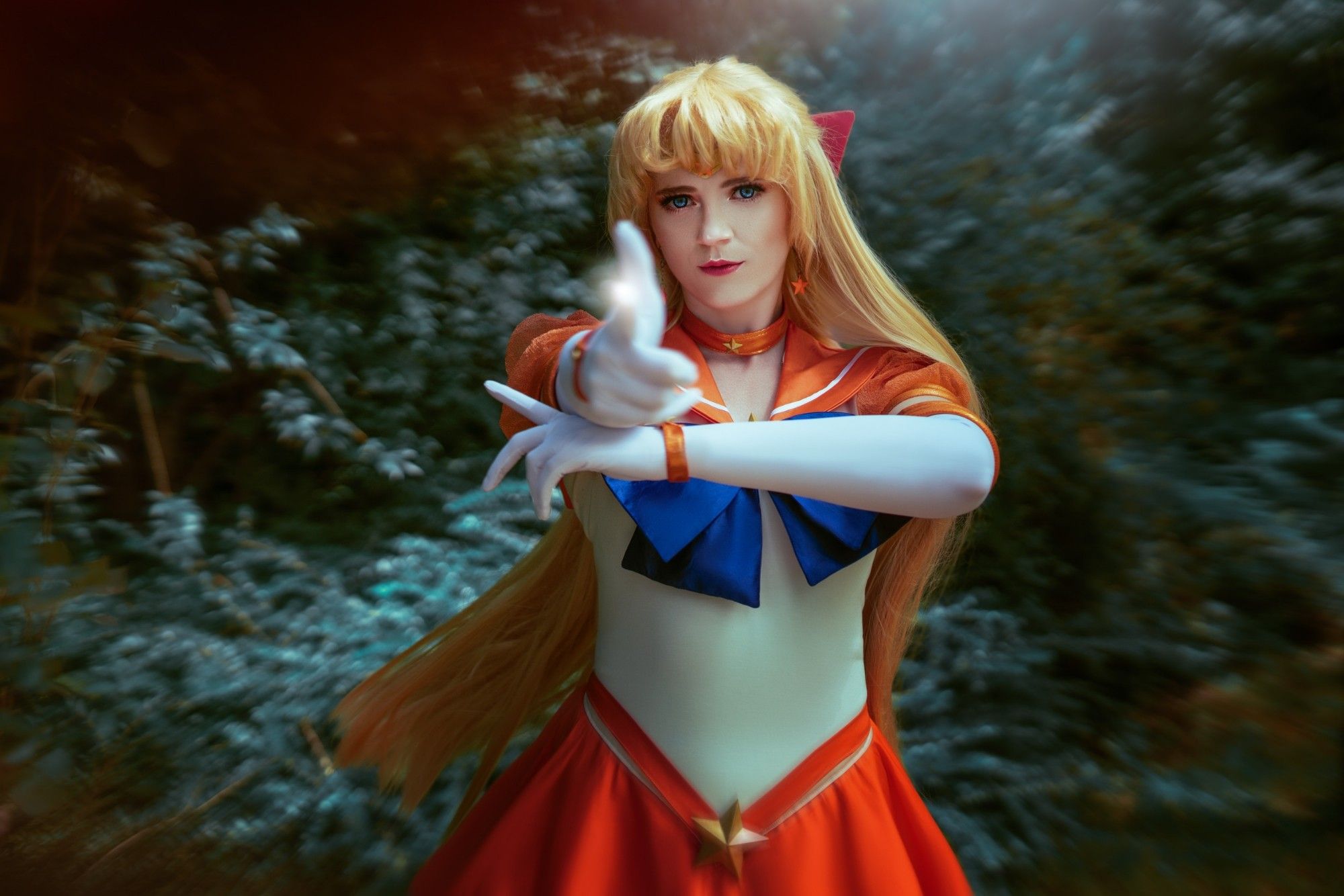 Female cosplayer as Eternal Sailor Venus with blond long wig and a white and orange suit, pointing with the finger in direction of the viewer, hair slightly swaying in the back, greenery as background