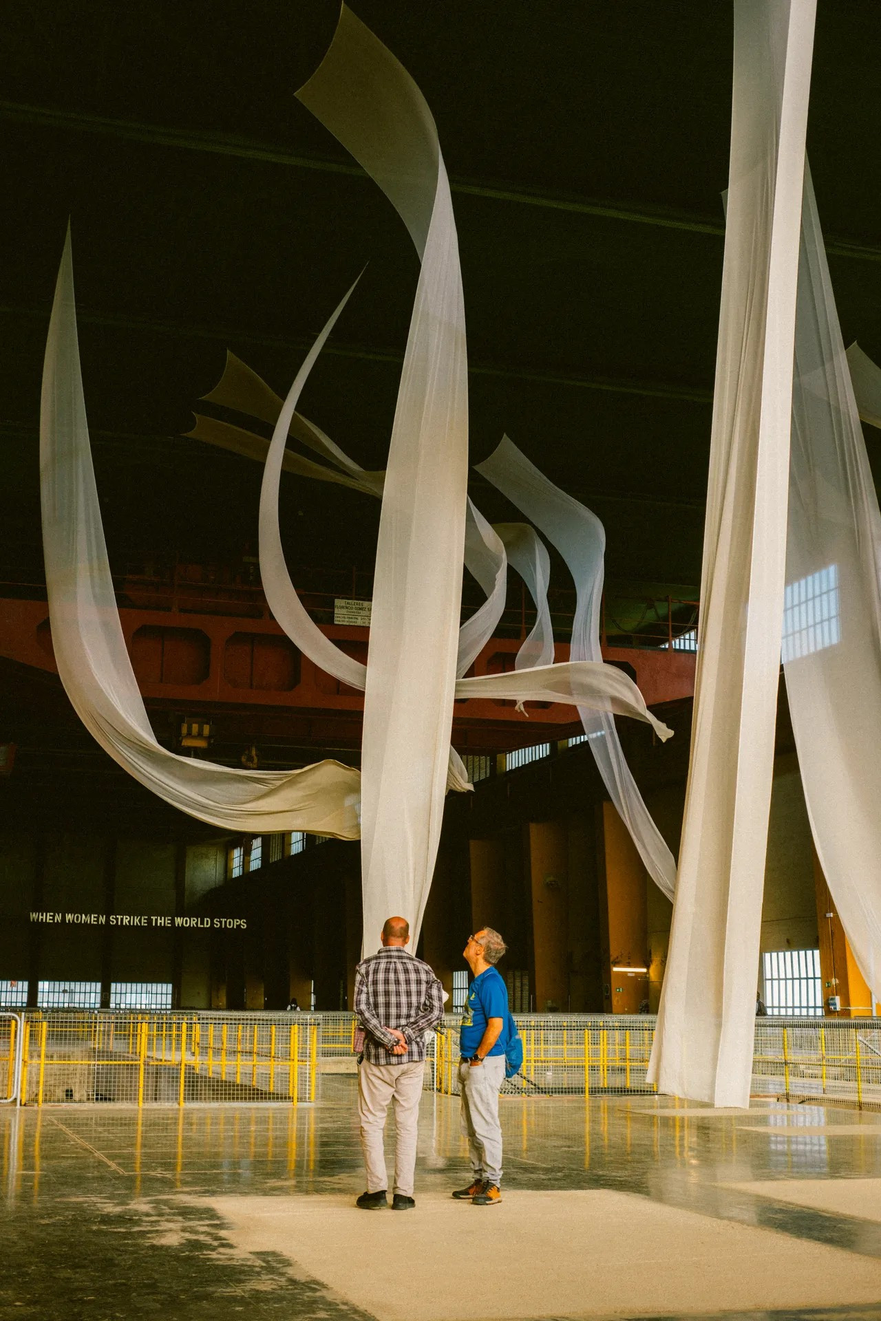 Dos hombres observan varias telas blancas suspendidas del techo en un gran espacio industrial. Al fondo hay un cartel que dice "When women strike, the world stops".