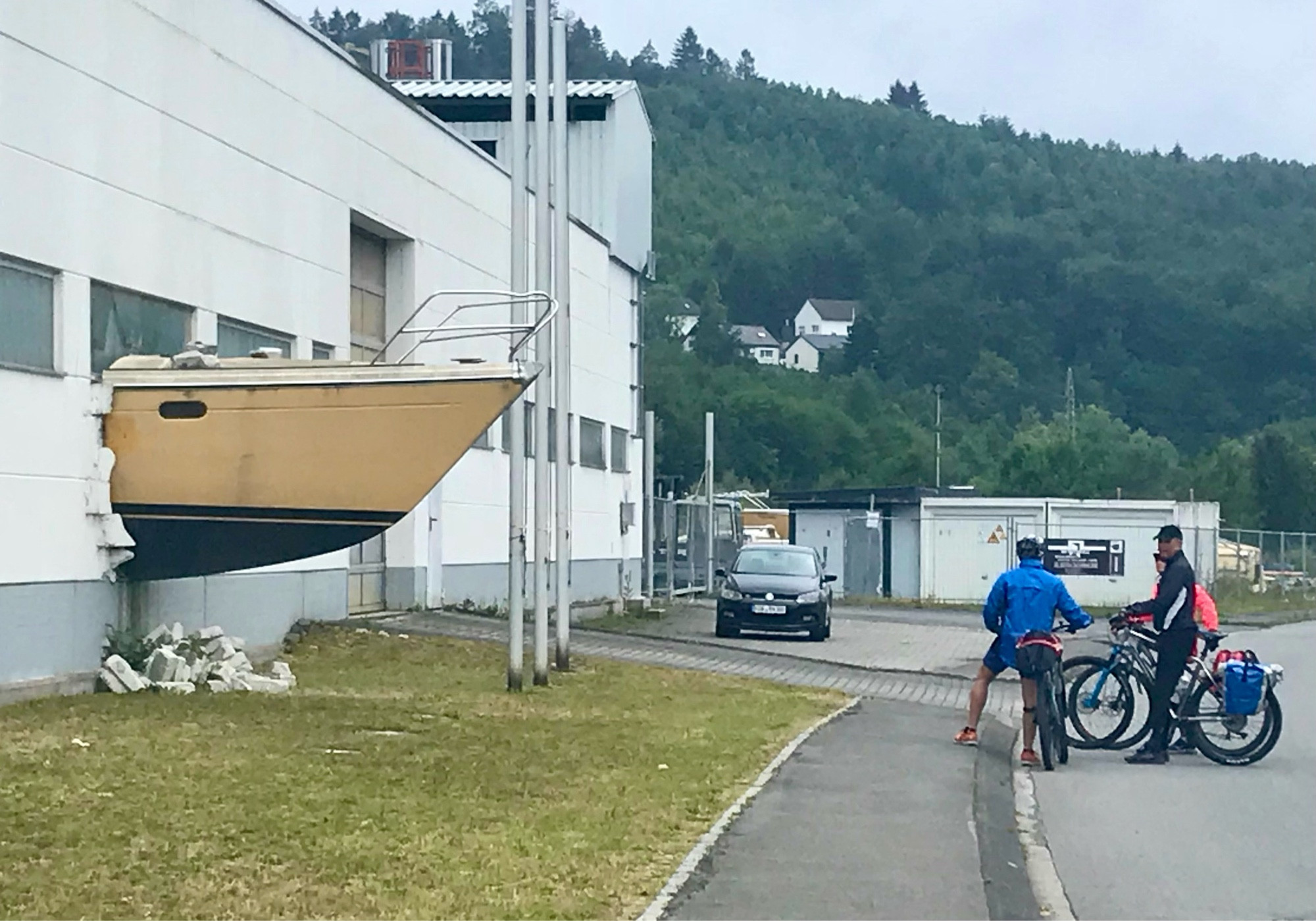 Ein Bug eines Bootes bricht scheinbar durch eine weiße Mauer. Vor der Mauer eine Wiese und ein Bürgersteig. Auf der Straße 3 
Menschen mit ihren Rädern.