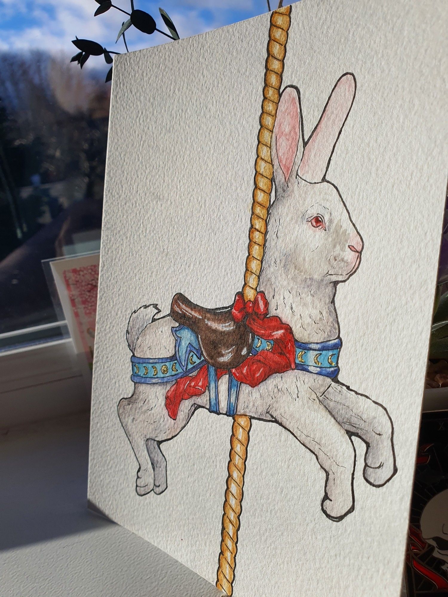 A watercolour painting of a white rabbit carousel animal. It has a brown saddle with a red bow ribbon rippling off it and blue attachments, which have golden moon phases decorating it. This photo is taken or the painting at a different angle to show the gold detailing more clearly.