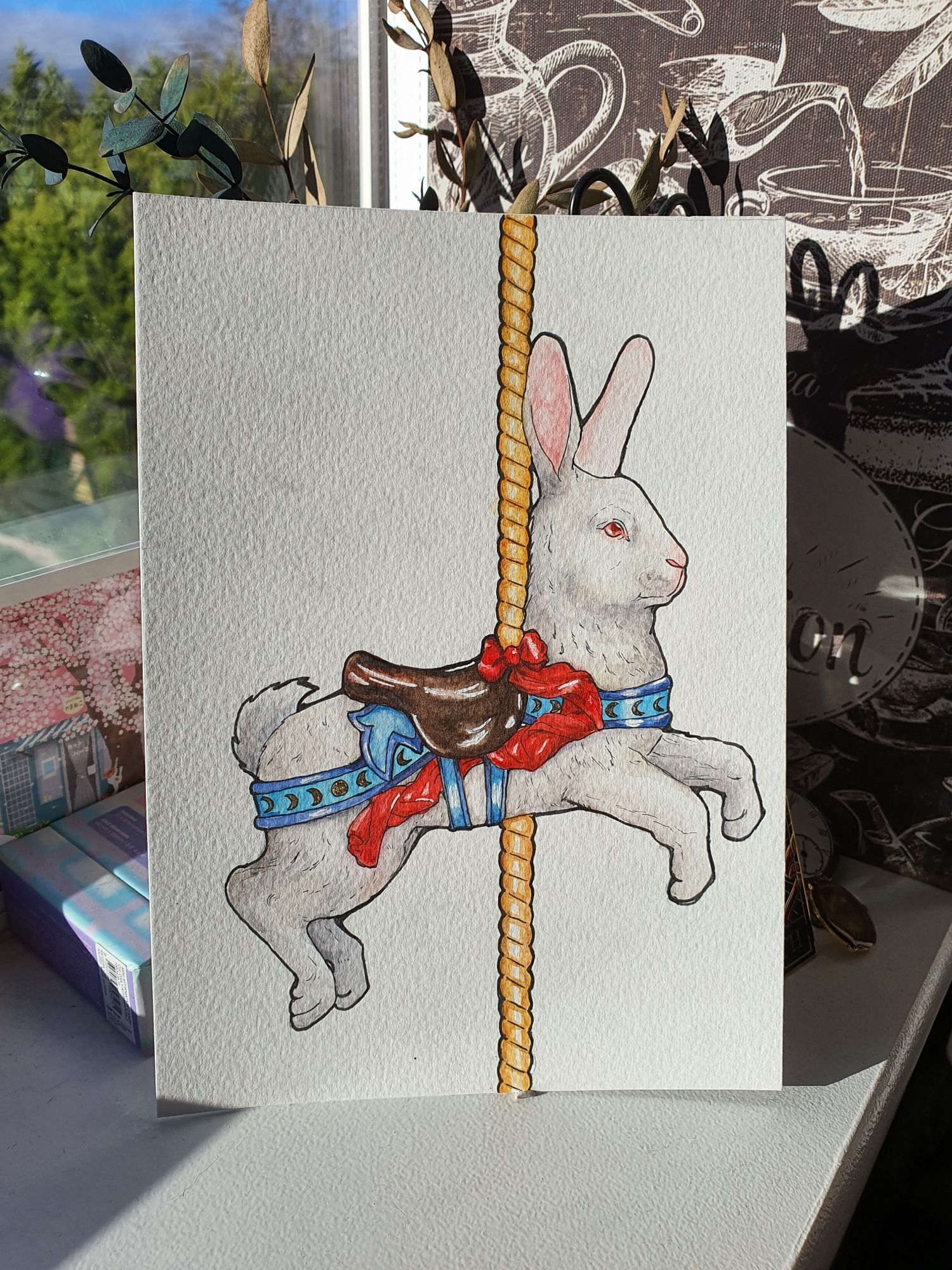 A watercolour painting of a white rabbit carousel animal. It has a brown saddle with a red bow ribbon rippling off it and blue attachments, which have golden moon phases decorating it.