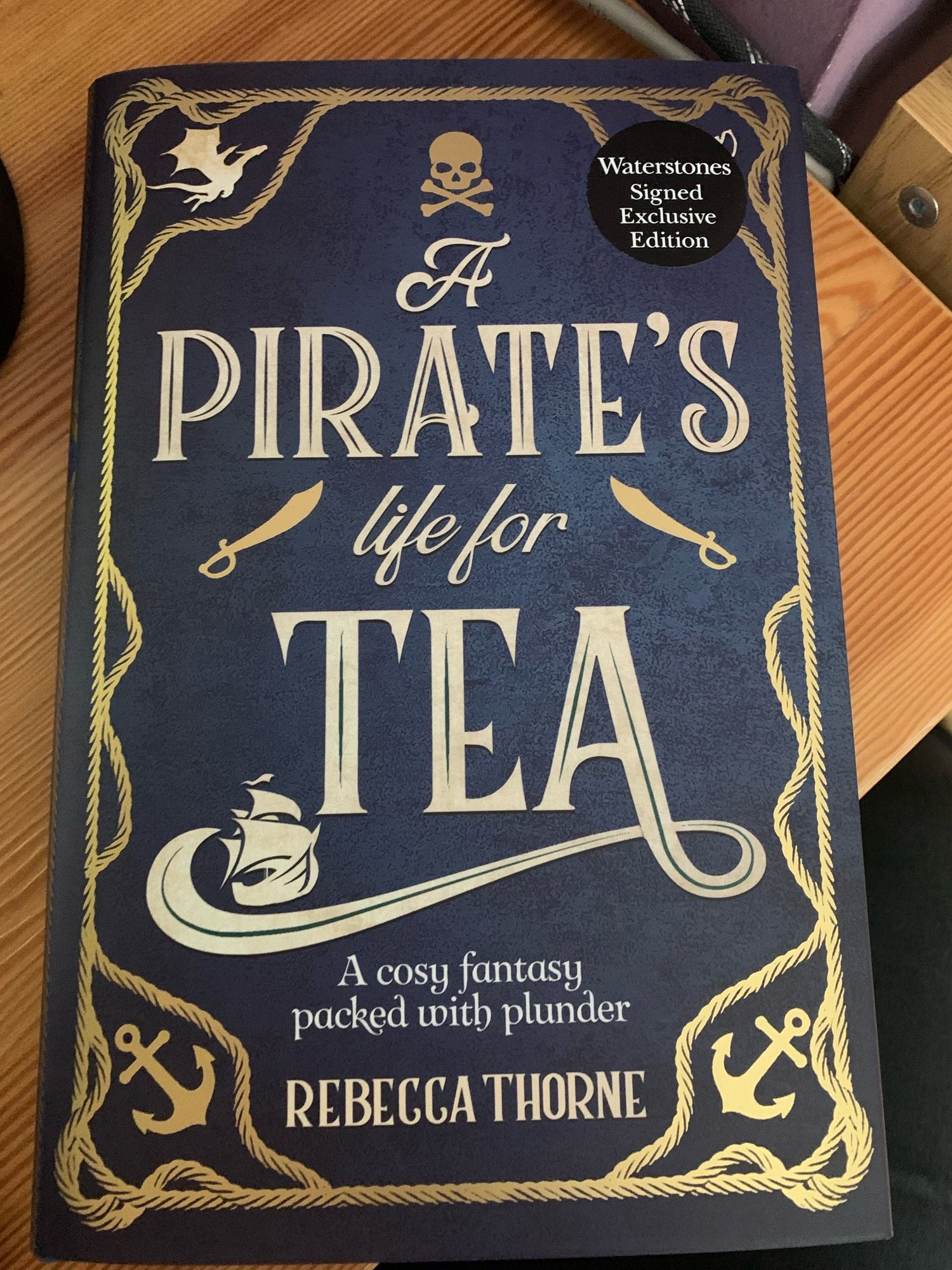 The front cover of A Pirate’s Life for Tea by Rebecca Thorne. The cover is blue with gold letting, gold rope design around the border with anchors at each bottom corner, a dragon in the top left, and whatever is under the top right is under a black Waterstones sticker which reads Waterstones signed exclusive edition. A skull and cross bones sits at the top, there is also a tall ship and a pair of cutlasses