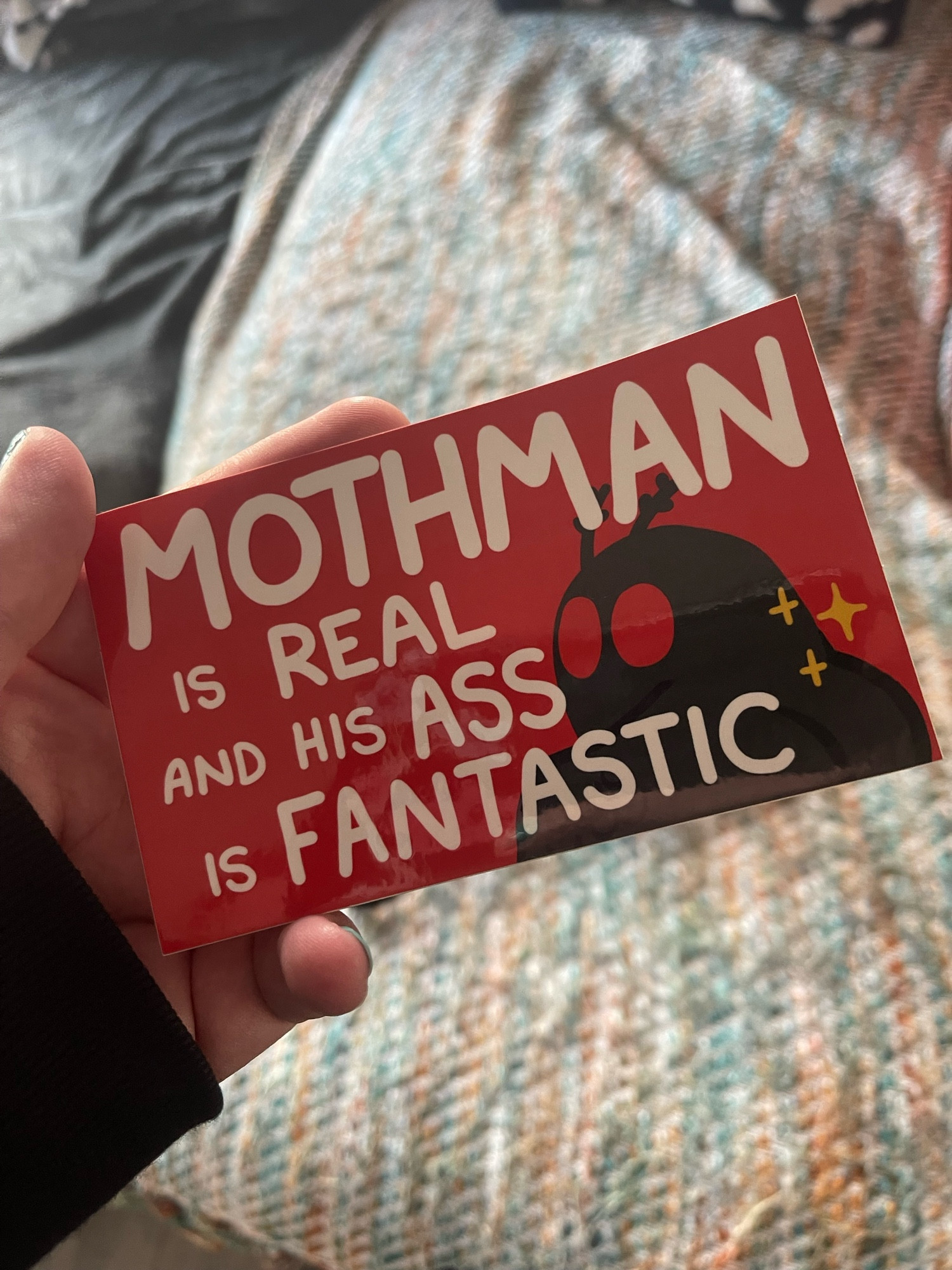 Riley holding a sticker that says “Mothman is real and his ass is fantastic” with an image of the cryptid on it