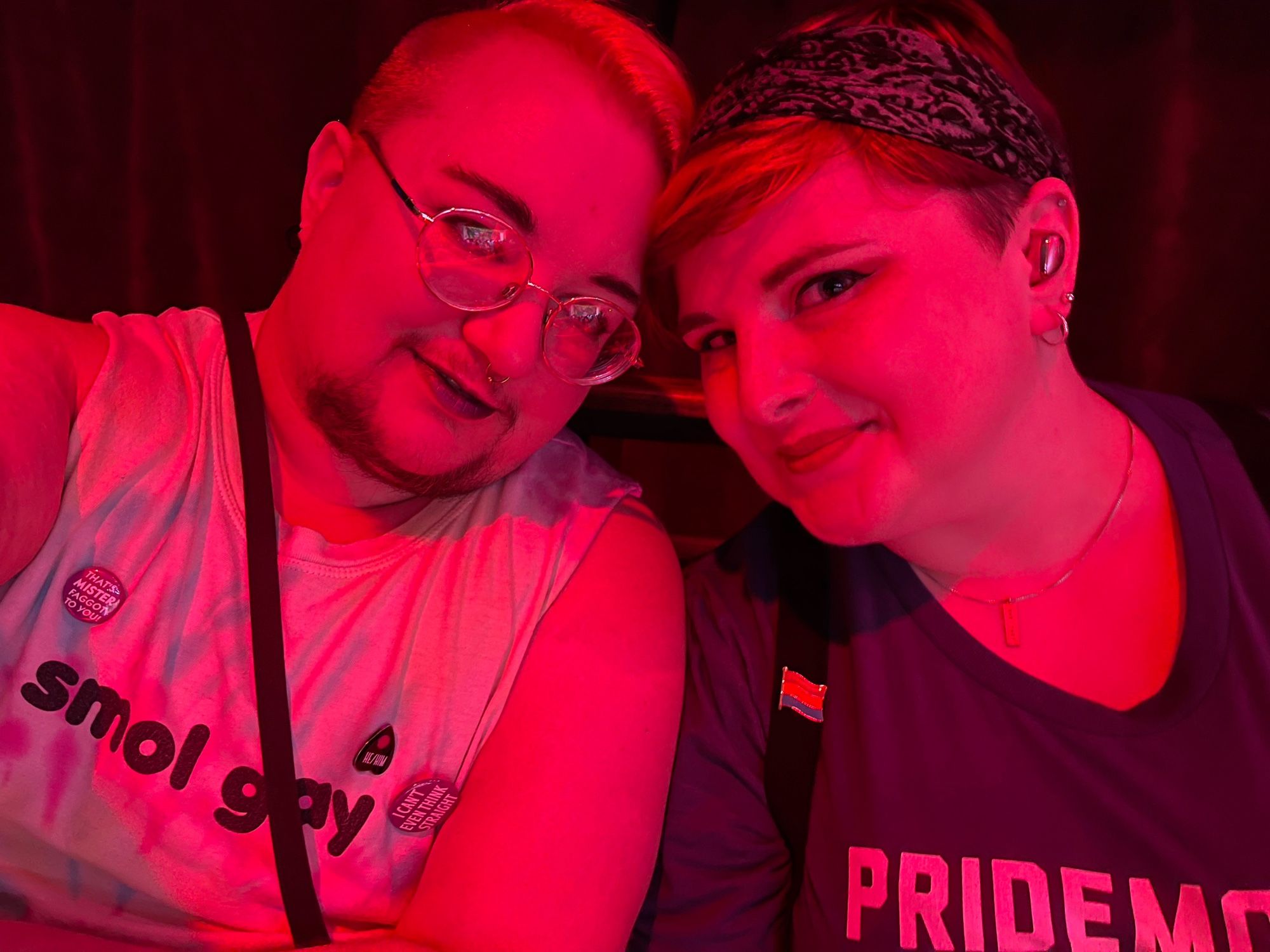 Two white trans people, one masc, one non-binary, in red-tinted light leaning their heads together. The masc one has a dark goatee and dark lipstick, the non-binary person has a pan pride pin and lighter lipstick. They’re wearing a shirt that says “Pride Month” that fades down to “Demon”, and the trans man is wearing a shirt that says “Smol Gay”. His pins say “he/him” on a planchette, “I can’t even think straight”, and “that’s MISTER faggot to you”.