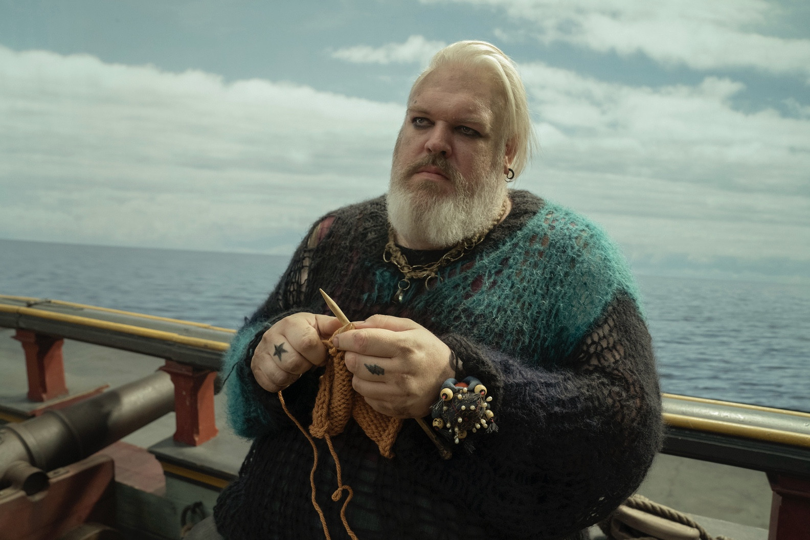 Kristian Nairn as Wee John Feeny, dressed in a green and black sweater with dark eye makeup. He leans against the railing of the Revenge while knitting.