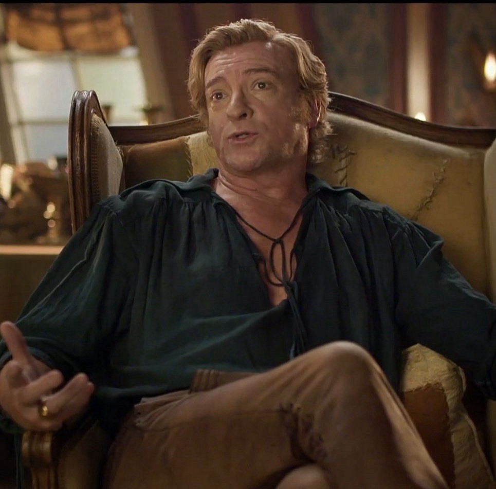 Rhys Darby in a behind-the-scenes clip, in full costume for Stede Bonnet. He wears tight brown trousers and a dark green pirate shirt with the tie open, exposing part of his chest. He sits with his legs crossed in a beige chair.