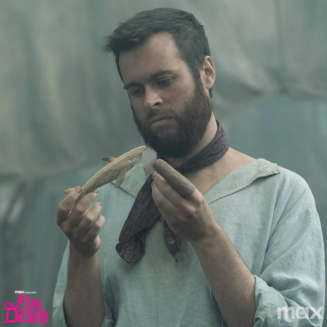 Nathan Foad as Lucius Soriggs holding a whittled shark. He has a dark scarf around his throat and is wearing a white shirt. He has a thick beard.