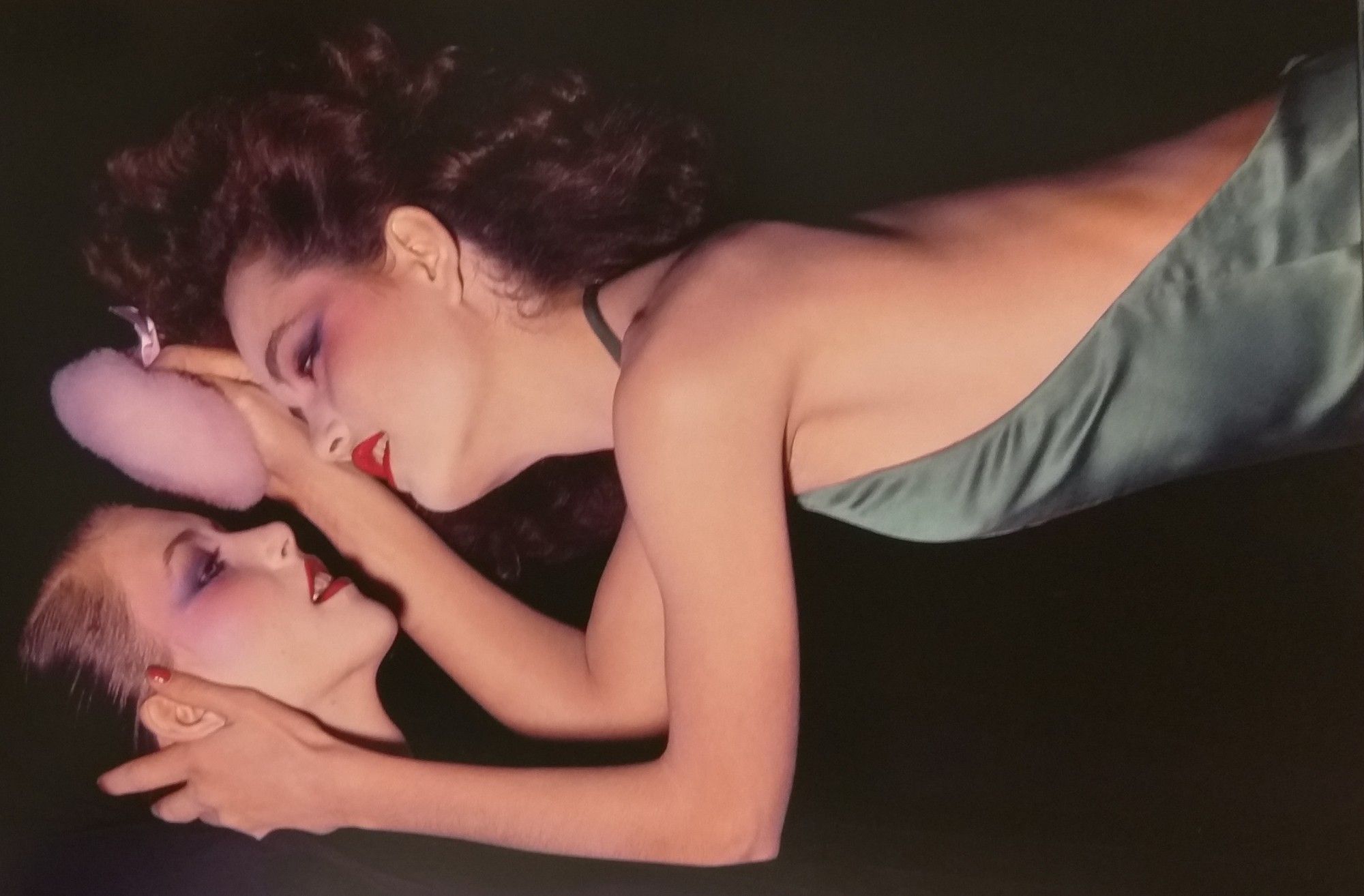 Foto: Guy Bourdin. imagem horizontal com fundo preto. Vemos a cabeça de uma mulher que está deitada, e uma outra mulher, usando um vestido frente-única de cetim verde, inclinada e prestes a se deitar sobre ela. ambas usam maquiagem sofisticada, com batom vermelho, blush e sombras nos olhos. Elas se olham e estão com as bocas entreabertas.