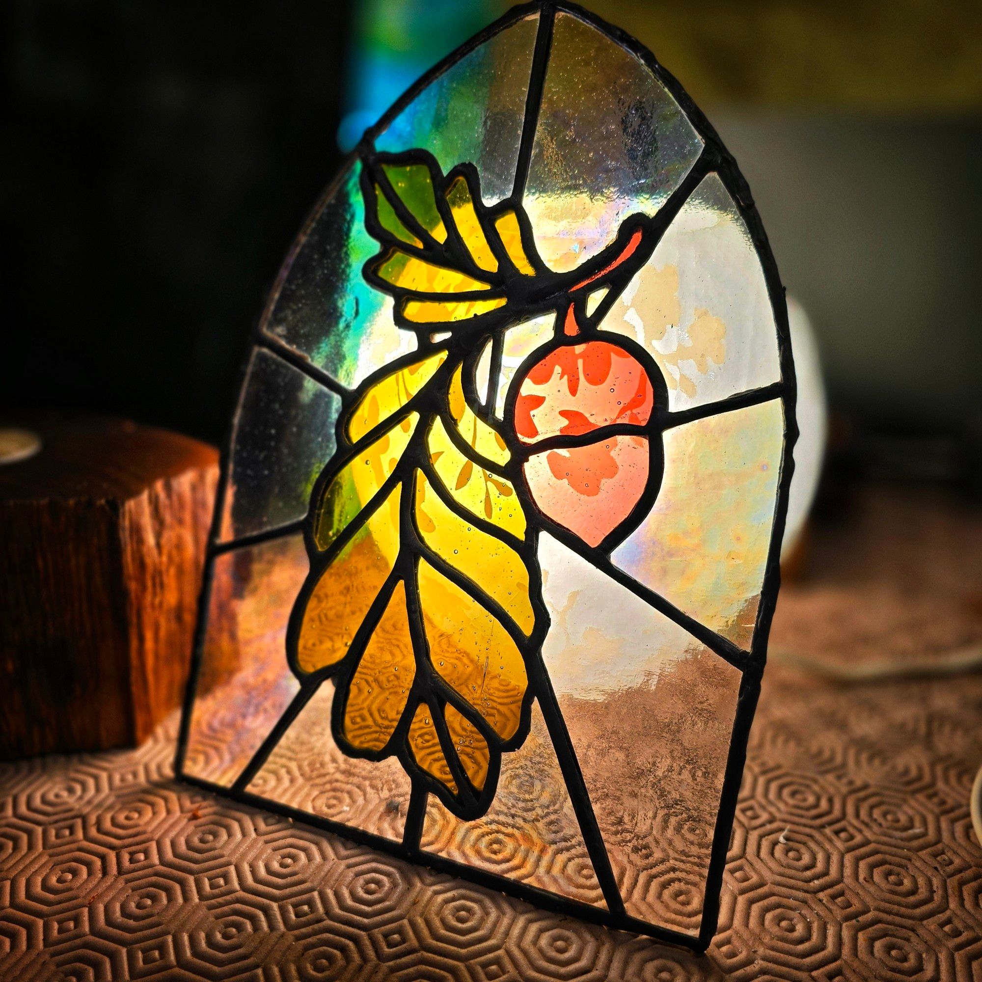 A stained glass artwork featuring a vibrant green-yellow oak leaf and a red-orange acorn hanging from a branch. The piece is framed in an arched shape, illuminated by warm light, highlighting the textured glass and rich colors. The background glass has an iridescent quality, casting soft reflections. It rests on a surface with a hexagonal pattern, adding a cozy, rustic feel to the scene.