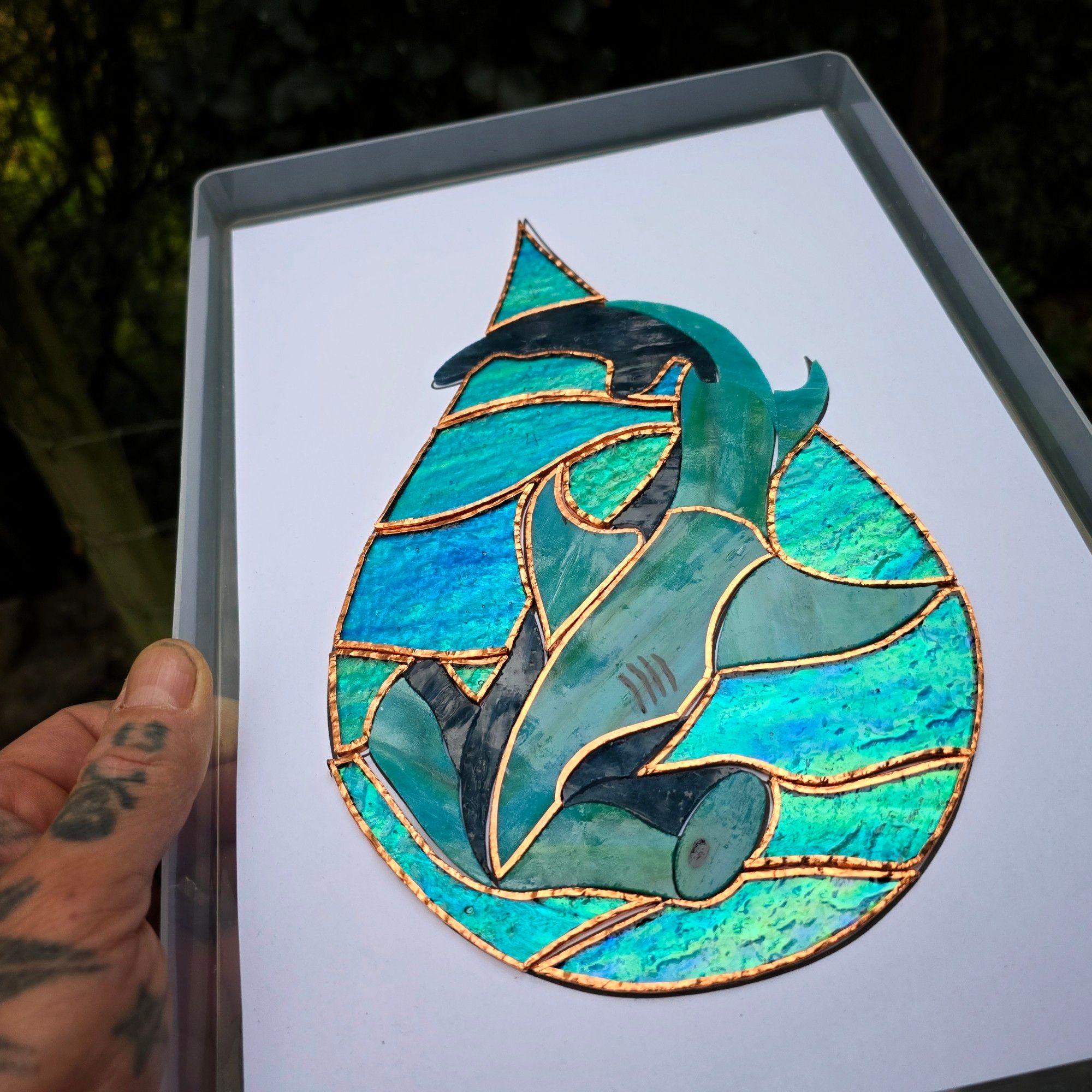 A nearly completed stained glass piece featuring a hammerhead shark, set against a watery blue background with copper foil outlining the sections. The shark is depicted in shades of green and gray, with the foil applied to most of the piece but a few areas still unfinished. The design has a flowing, circular composition, mimicking the movement of water. The piece is shown on a flat surface in natural light.