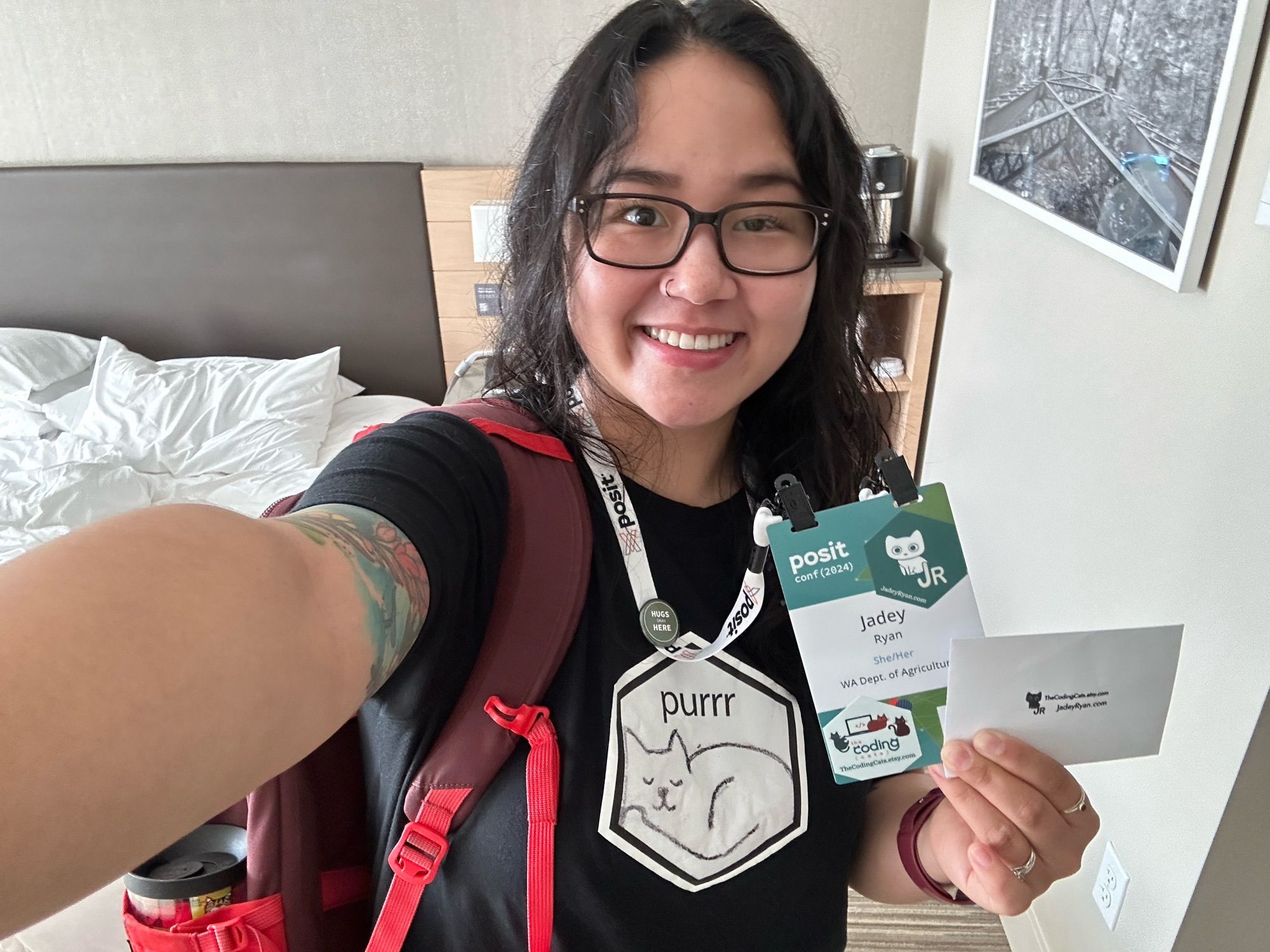 Jadey smiling wearing a purrr tshirt and red backpack. She’s holding up her posit conf badge with her hex stickers and envelope for safely storing all the stickers!