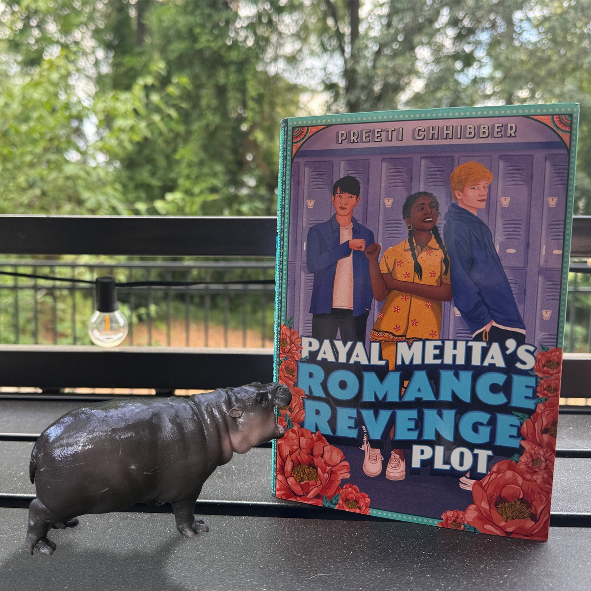 A copy of PAYAL MEHTA'S ROMANCE REVENGE PLOT sitting on a table with the pygmy hippo Moo Deng next to it, trying to bite the cover.