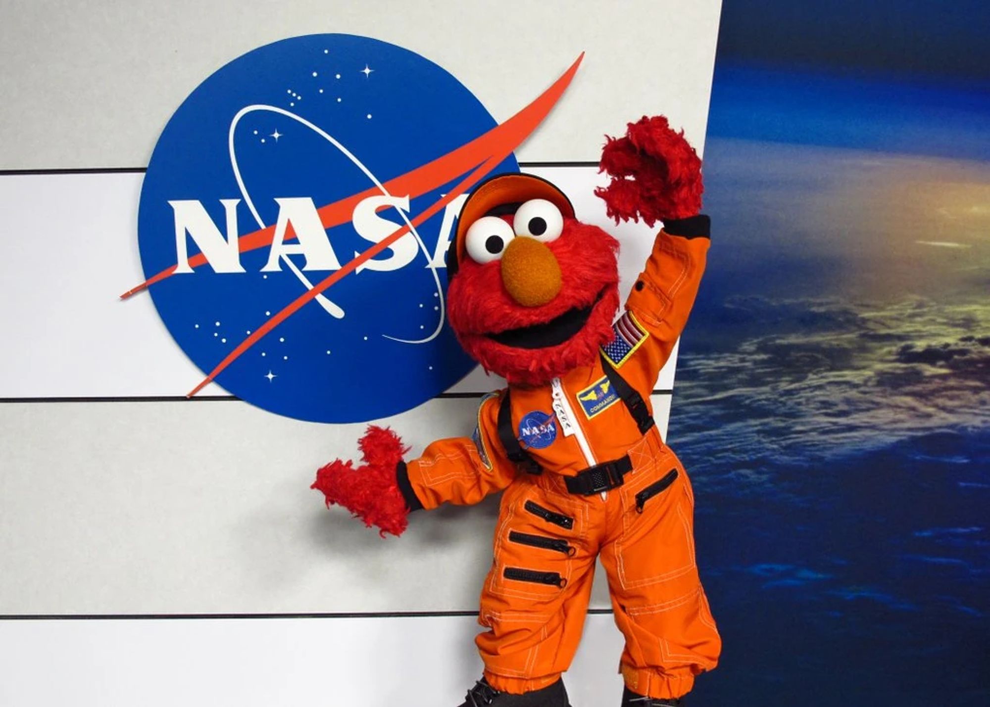 ID: Elmo stands in an orange NASA space suit and hat by the NASA logo with an image of earth from space in the background to the right.