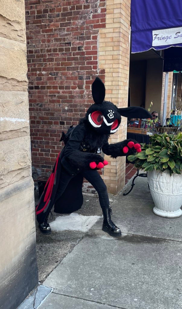 Black moth fursuit named Moff with red eyes and white under eye markings. He has a white eye marking on the forehead. Moff is peaking out of an alleyway at a festival. 