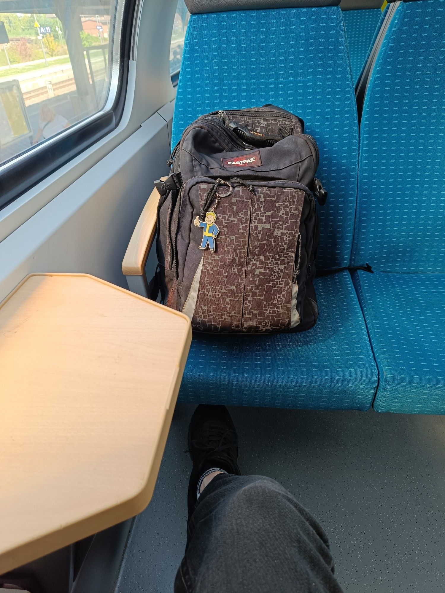 Ein Vierer-Sitzbereich eines Regionalexpress es.
Blau bezogene Sitze.
Links im Bild Fenster und kleines Tischchen.
Im gegenüber liegenden Sitz am Fenster ein Eastpak-Rucksack. An einem der vorderen Reißverschlüsse baumelt ein Vaultboy-Schlüsselanhänger.
Ein Vierer-Sitzbereich eines Regionalexpress es.
Blau bezogene Sitze.
Links im Bild Fenster und kleines Tischchen.
Im gegenüber liegenden Sitz am Fenster ein Eastpak-Rucksack. An einem der vorderen Reißverschlüsse baumelt ein Vaultboy-Schlüsselanhänger. 
Von unten ragt ein Bein in einer schwarzen Jeanshose ins Bild.