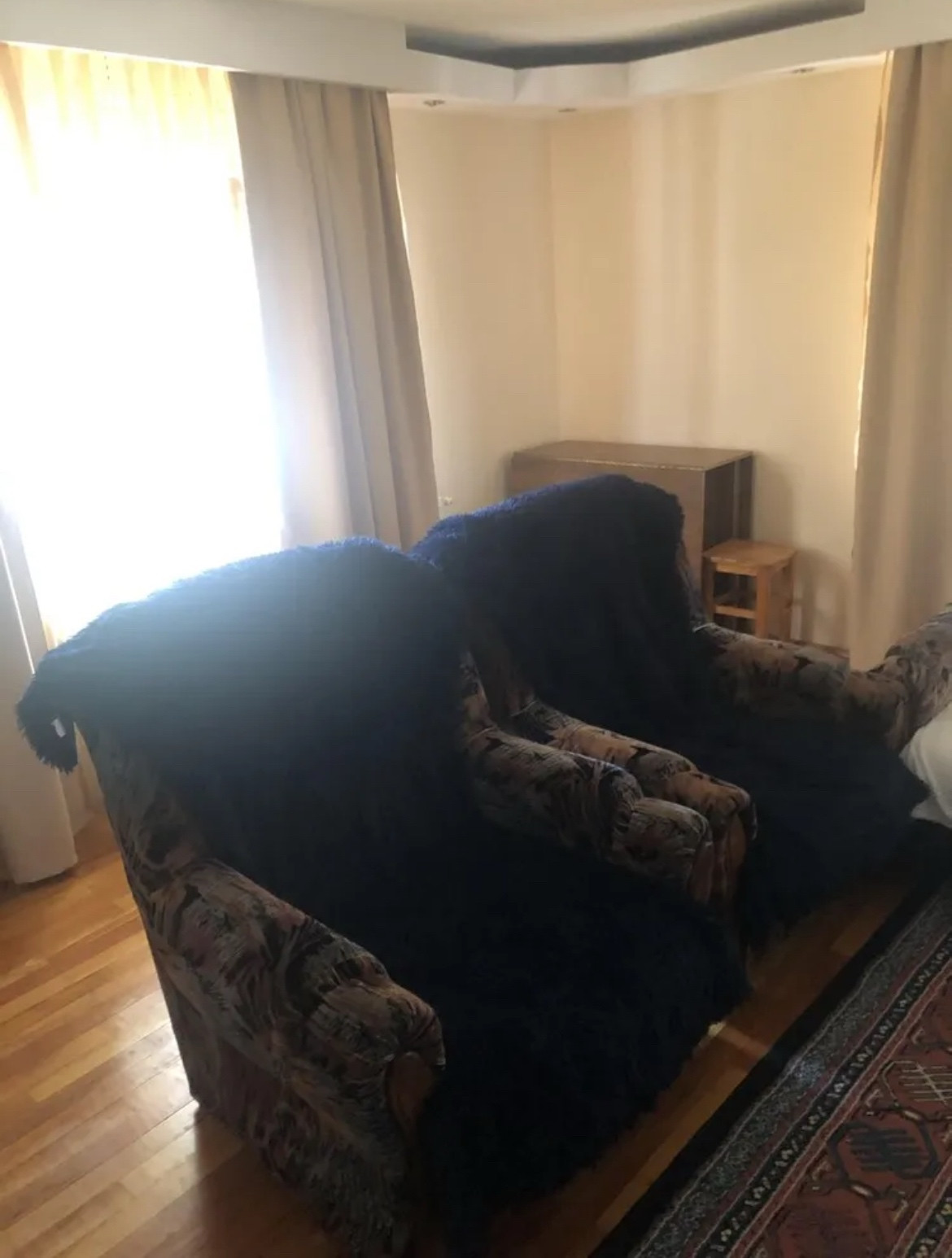 Verschwommenes Fotos in ein Wohnzimmer. Präsent sind zwei dunkel gemusterte große Sessel die nebeneinander stehen, dahinter eine wand mit durchsichtigen Vorhängen, denen sich ab den seiten eines erahnbaren Fensters noch dunkle Vorhänge anschließen. Außerdem hat eine graue Kommode hinter den Sesseln in der Zimmerecke ein Plätzchen gefunden. Der raum sieht aus als würde er muffig, staubig und nach alten menschen riechen.