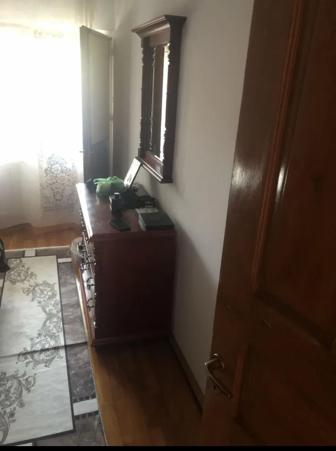 Womöglich der andere Teil des Wojnzimmers. Rechts im Bild ist eine dunkle Eichentür, gefolgt von einer beigen Wand an der eine Kommode Typ Eiche rustikal steht. Auf der Kommode stehen dunkle Dekogegenstände über der Kommode hängt ein großer Eiche rustikal Bilderrahmen.Abschluss der Wand wieder eine halboffene Tür vor hellen Vorhängen. Auf den Boden liegt ein Teppich der eine starke Falte von quadratisch zusammenfalten aufweist. 