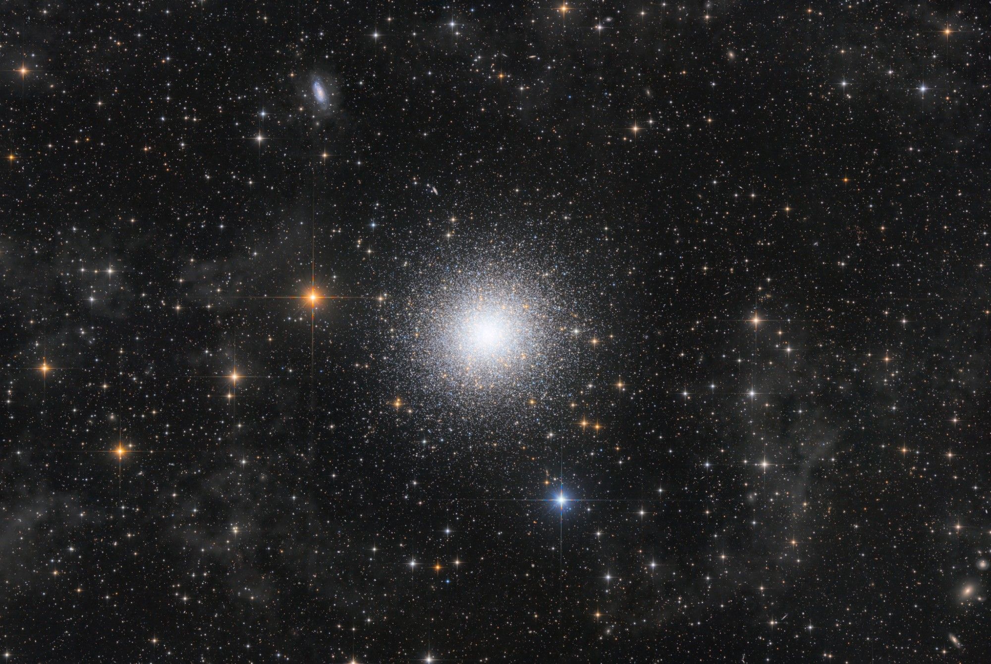 astrophotograph of a very rich star field. In its centre, is a spherical shaped ball of many stars, all clustered together. This is the globular cluster. It has very bright white stars, peppered with orange and yellow stars too. Surrounding it are soft low density clouds of gas, and off in the distance are several galaxies. The brightest stars in the image have diffraction spikes.