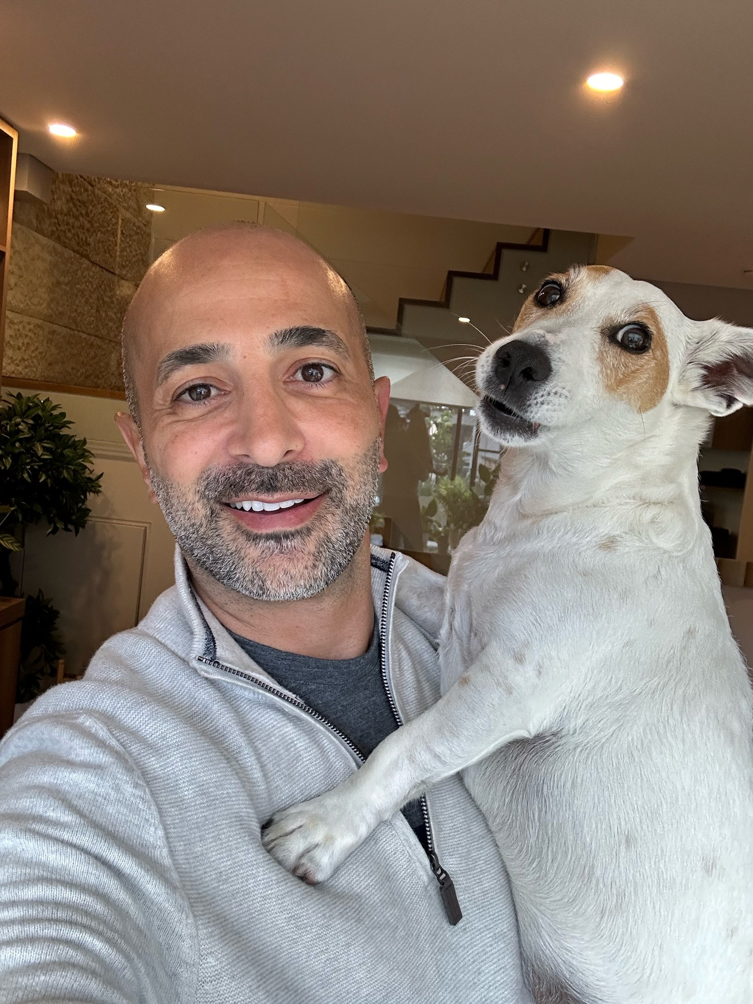 A man taking a selfie and he is holding up a small white dog, who is also looking at the camera in a goofy manner. The dog is perched up on the man’s shoulders supporting itself with its front paws. 