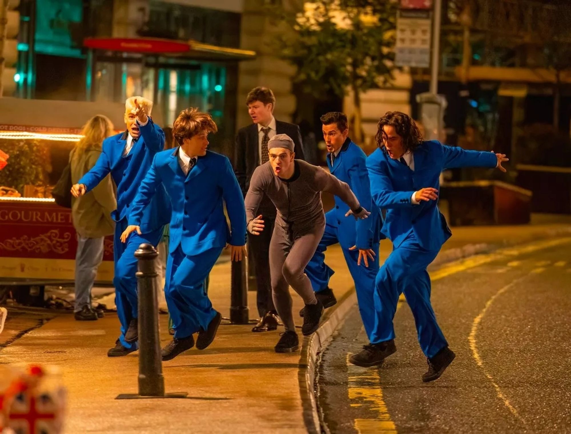 A man in a grey bodysuit for CGI purposes surrounded by a group of men in blue suits depicting a young Take That running down Regents Street, London