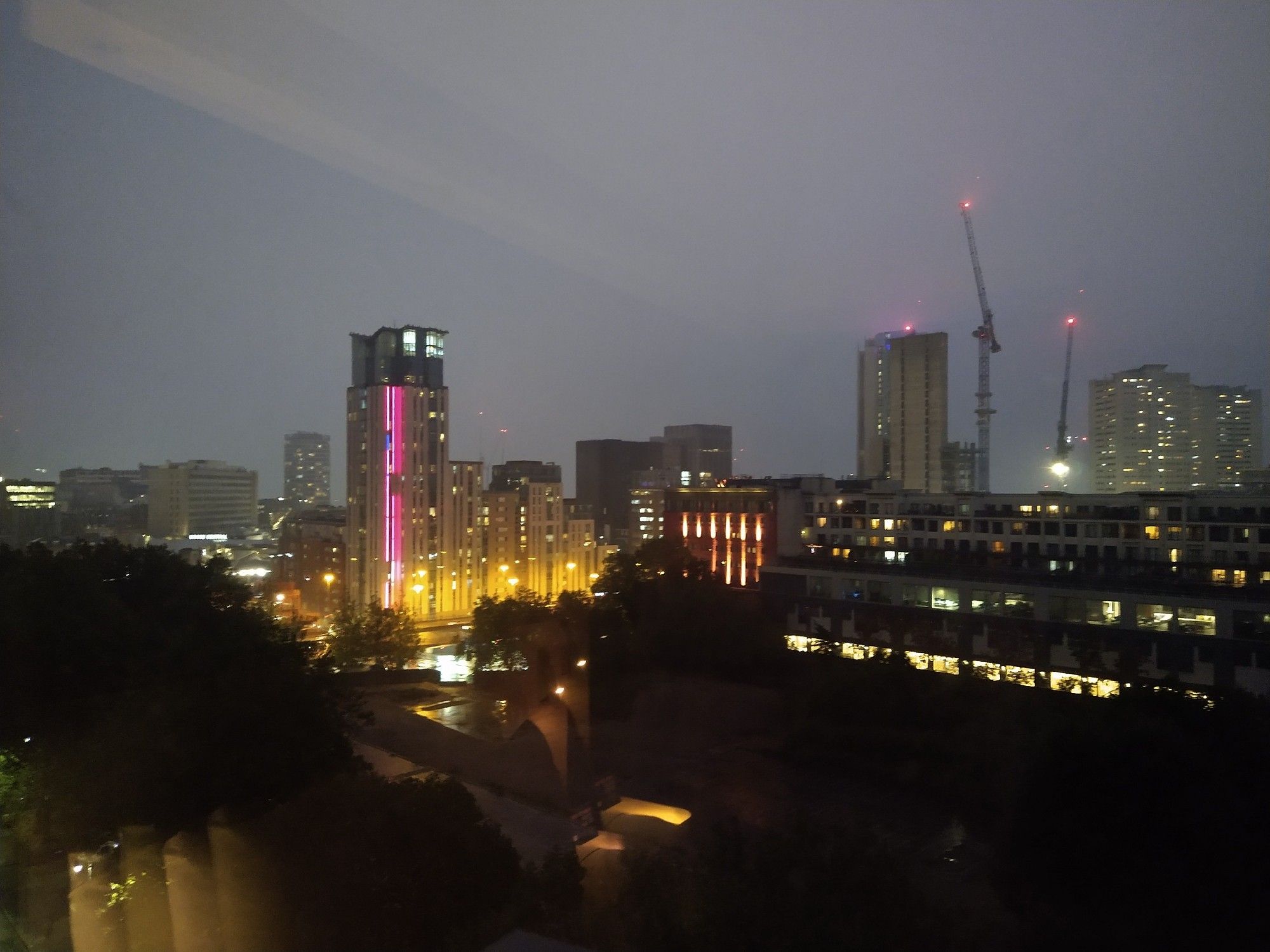 A grey and rainy dusk falls over Birmingham - a collection of modern high rises and construction cranes lit in neon pinks and reds.