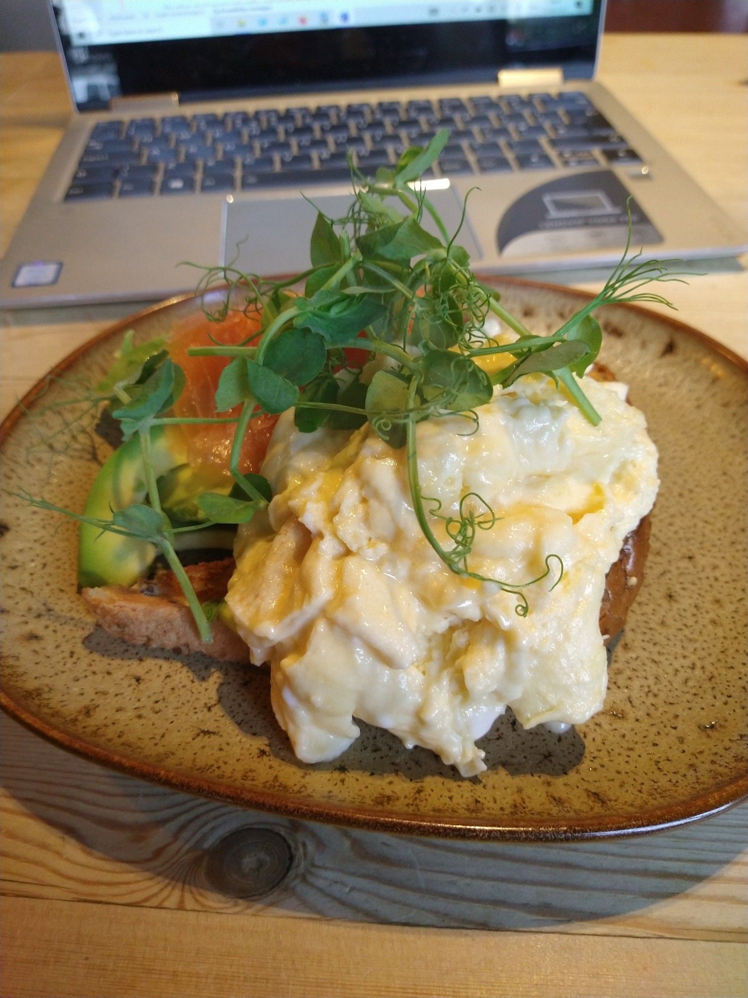 A close up of scrambled eggs and smoked salmon on toast, with a laptop computer open in the background.