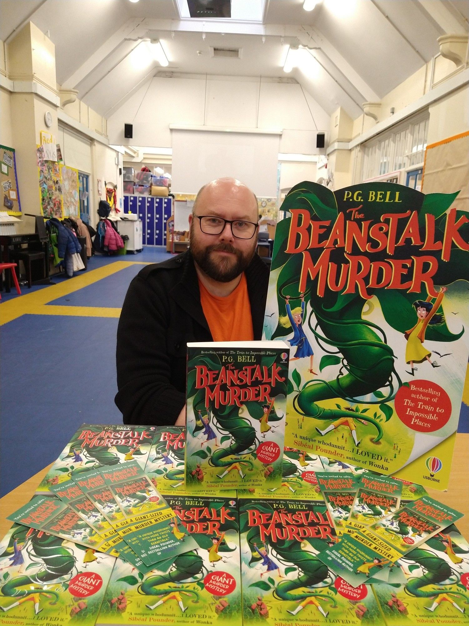 P.G. Bell sitting at a table piled high with copies of The Beanstalk Murder, in a primary school hall.