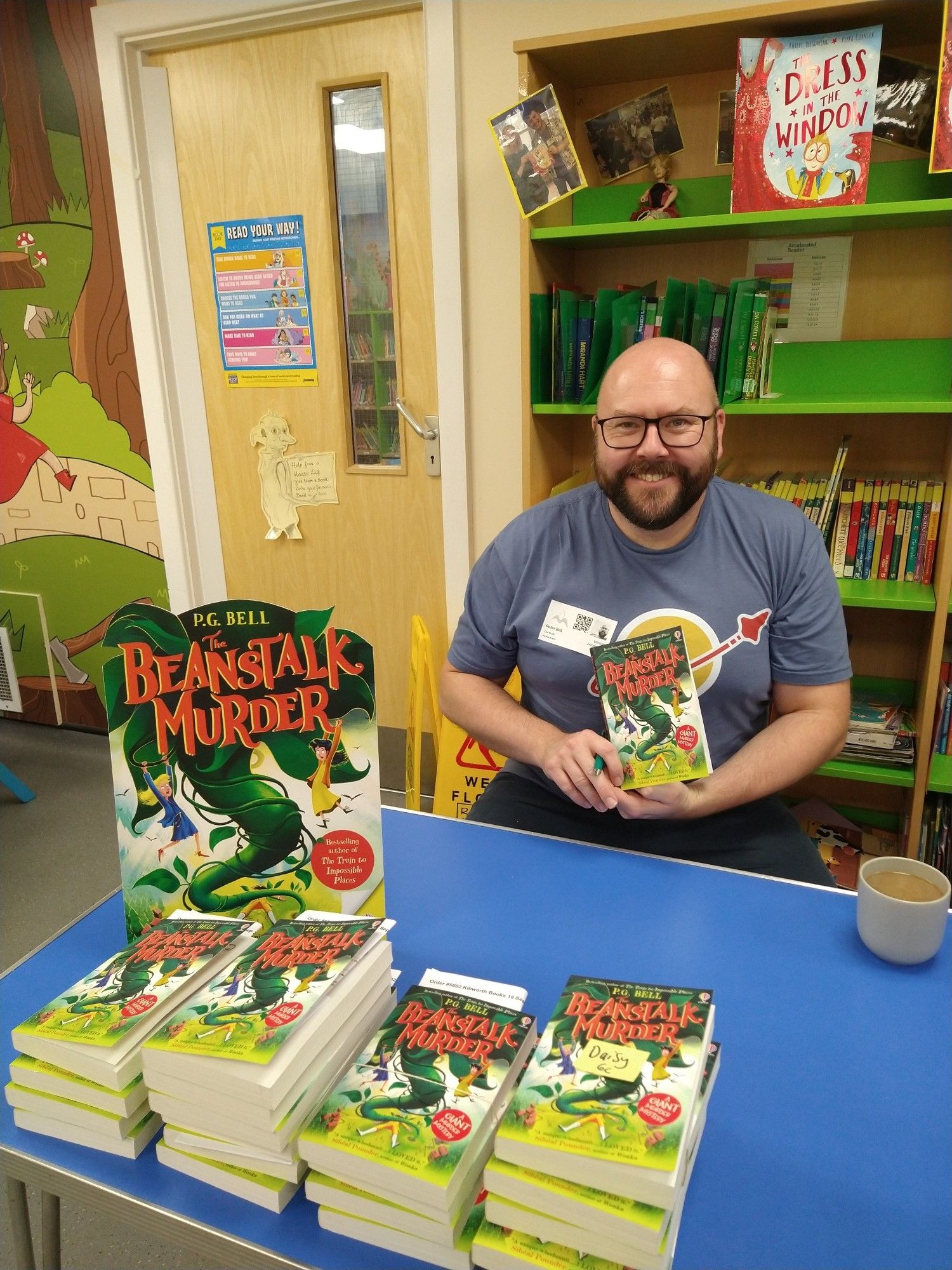 The author P.G. Bell sitting at a table in a primary school library, behind stacks of copies of The Beanstalk Murder and a cup of (very welcome) coffee.