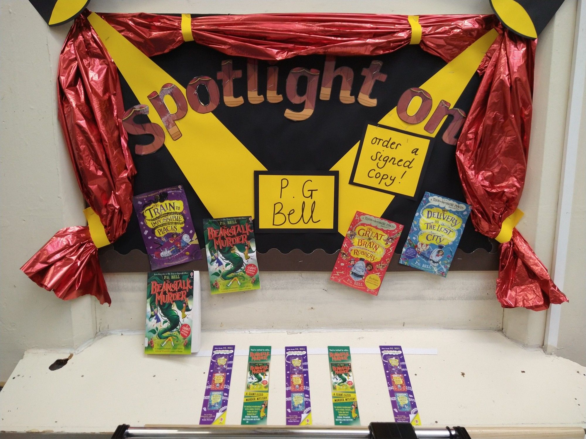 A wall display of P G. Bell's books, under the words "Spotlight On". The display is framed by red theatre style curtains and yellow cardboard spotlights.