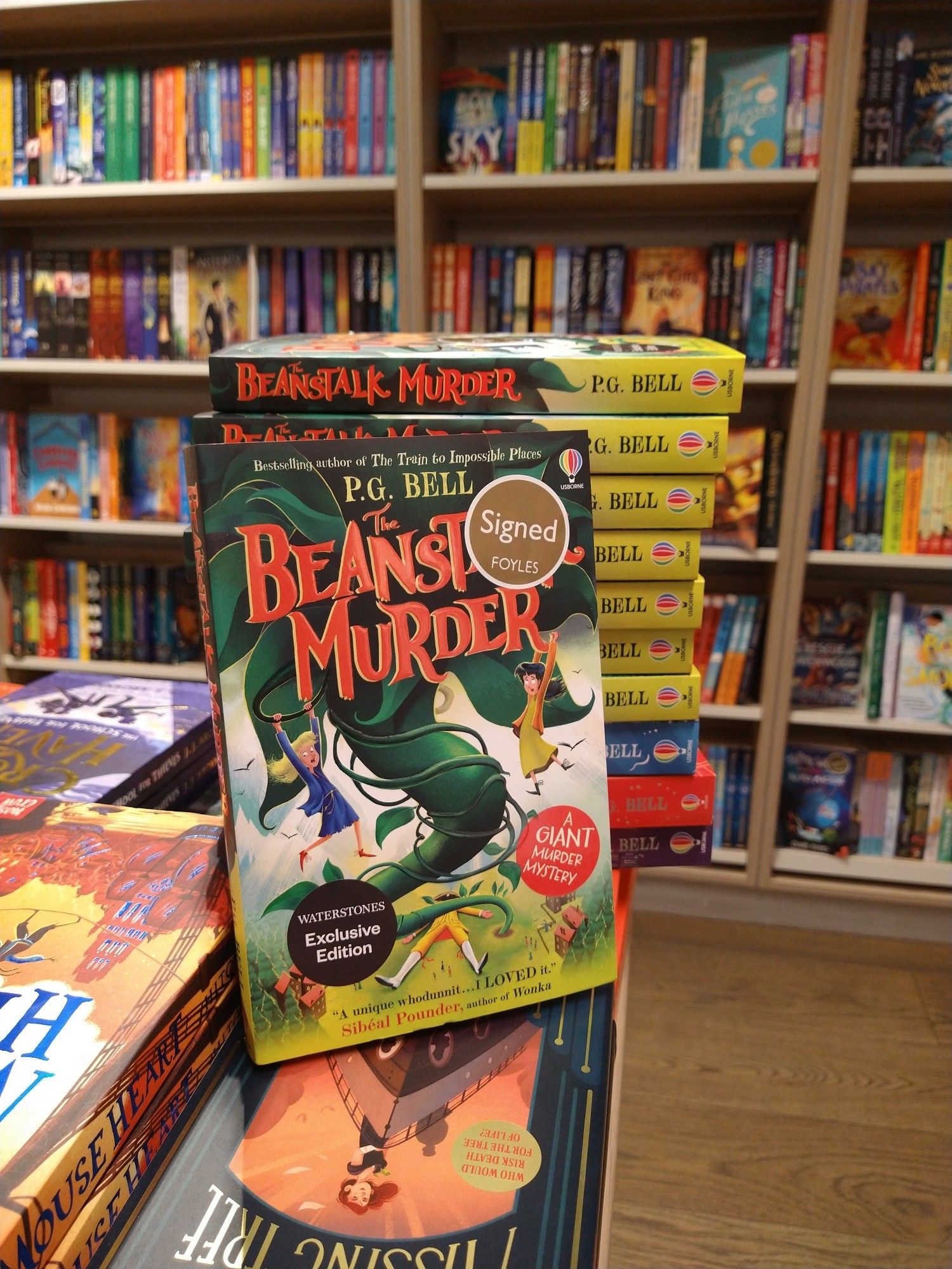 A stack of copies of The Beanstalk Murder paperback. The cover features two young girls hanging from a giant beanstalk high above a country village.