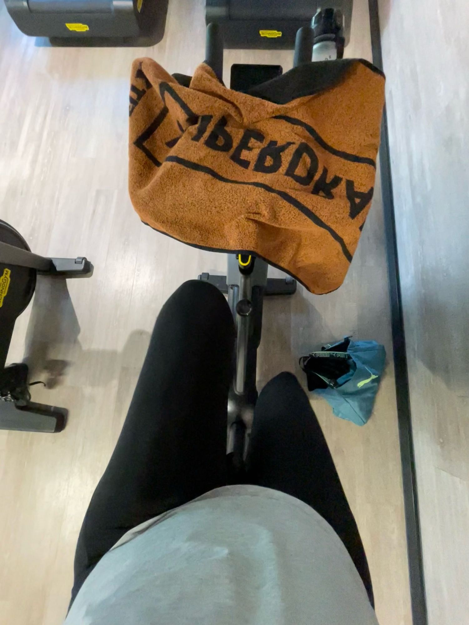 POV, ich sitze auf dem Rad und schaue runter auf meine Beine. Man sieht eine schwarze sportleggings, ein graues top, ein orange-schwarzes Handtuch auf dem Lenker, die sporttasche ist petrol und liegt neben dem bike auf dem Boden