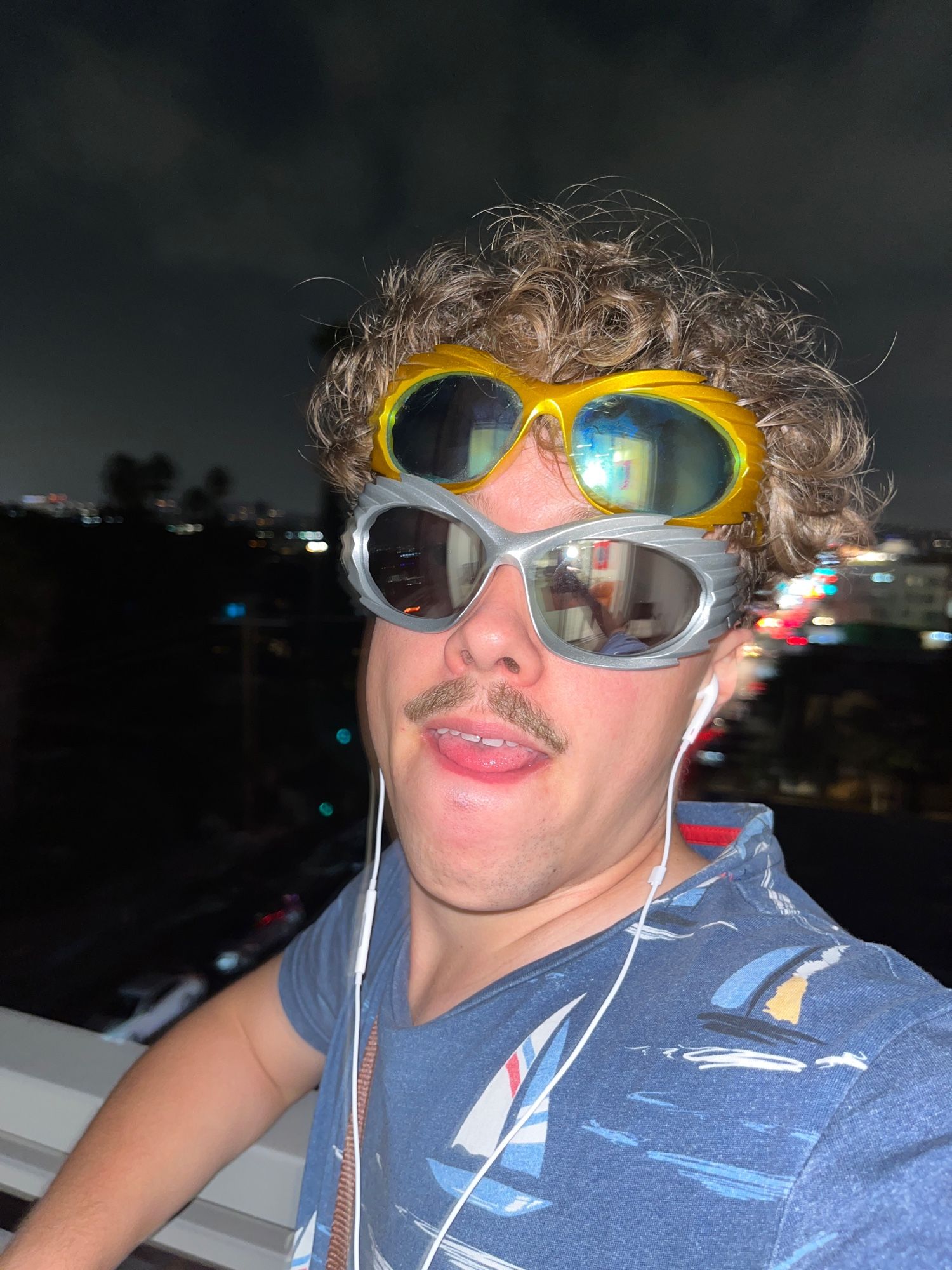 Crazy cool guy wearing multiple raver sunglasses with white in-ear headphones on a balkony in Los Angeles. He has cool boats on his blue T-Shirt.