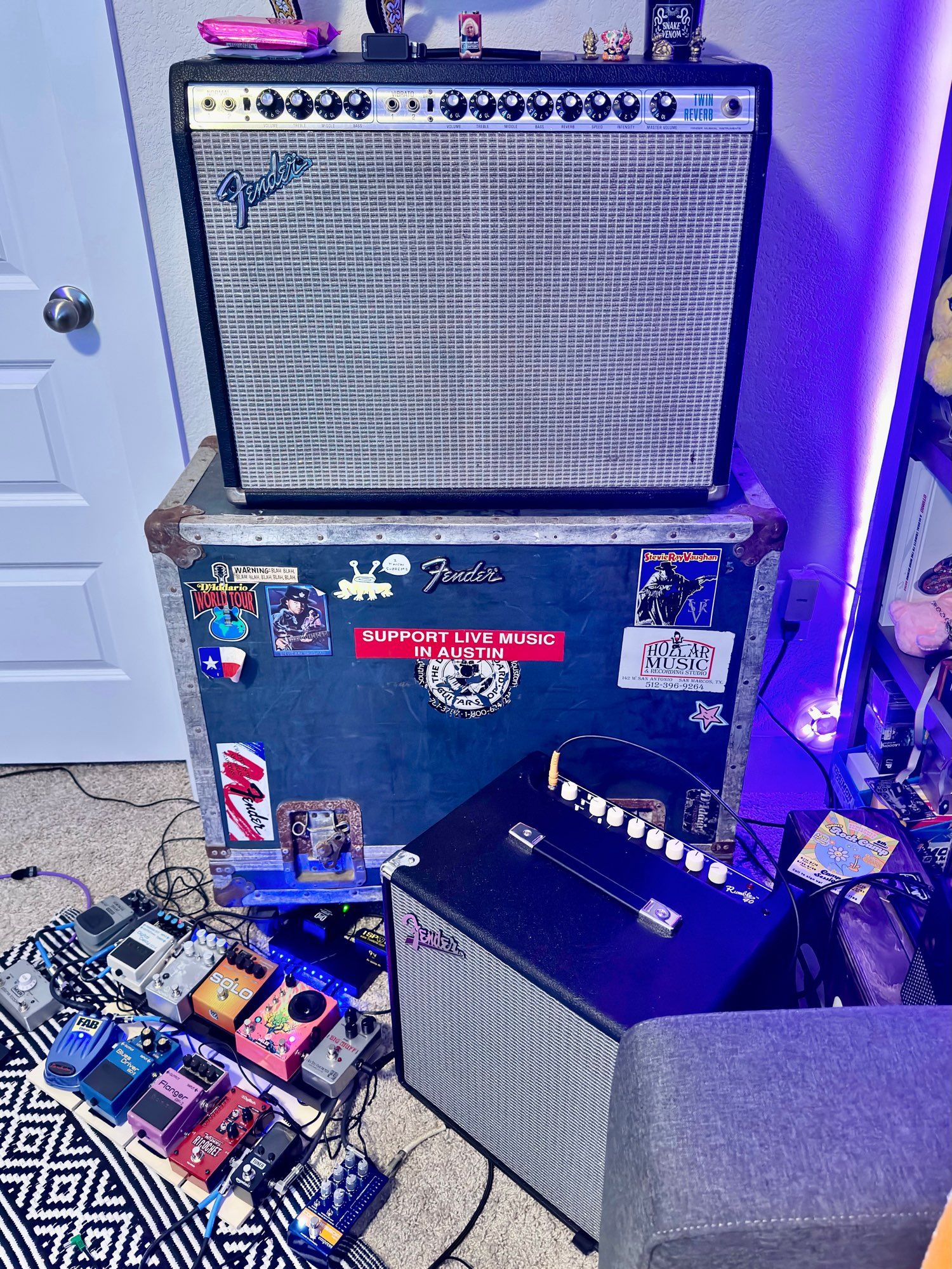 A twin reverb sitting on top of a matching era road case. Red bumper sticker reads “support live music in Austin” and there are several old Stevie ray stickers and vintage fender bumper stickers. Rusted edges. Pedal board sitting at the base of the stack.