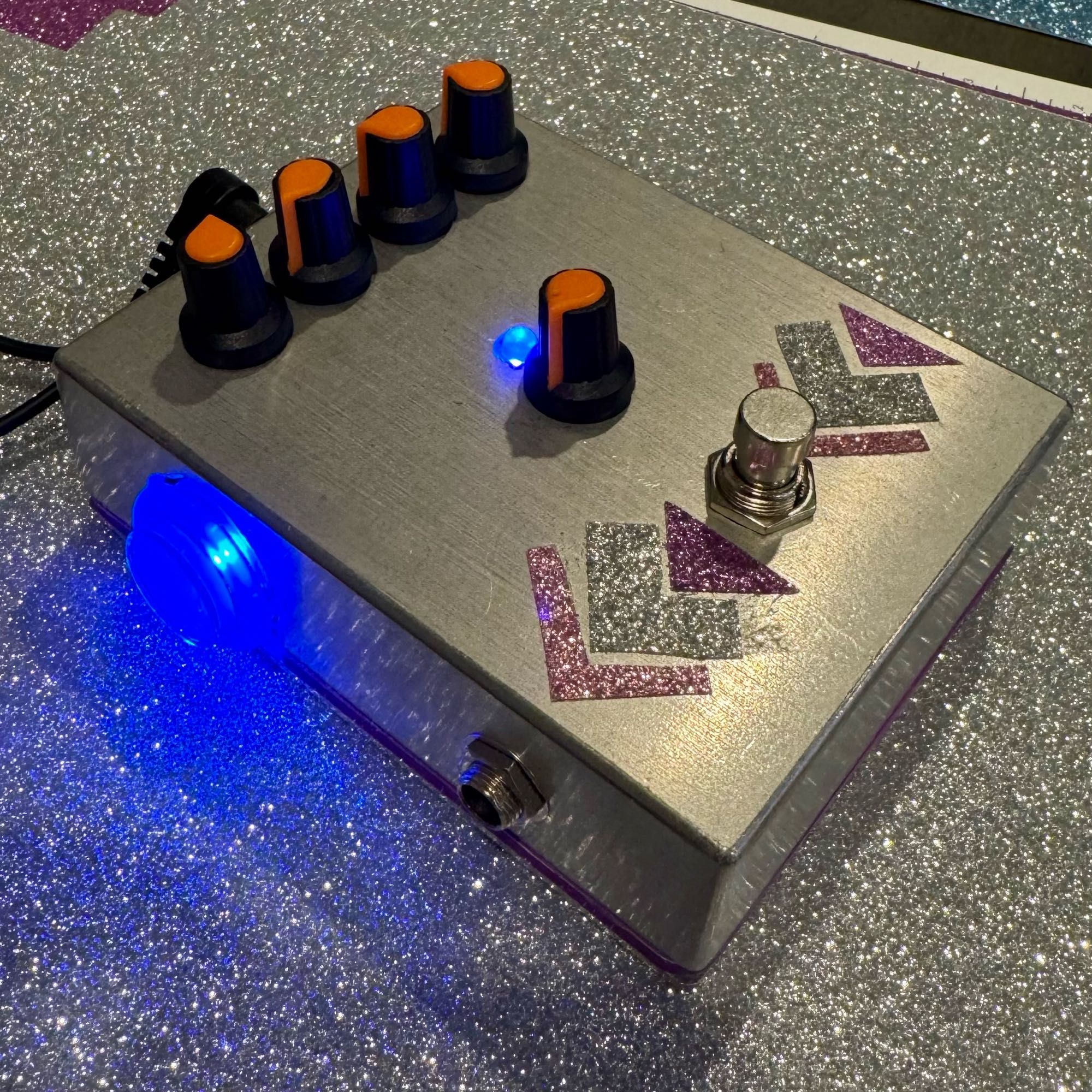 Photo of the finished pedal. Four knobs on top. Arcade button lit up on the left and a center knob for main volume