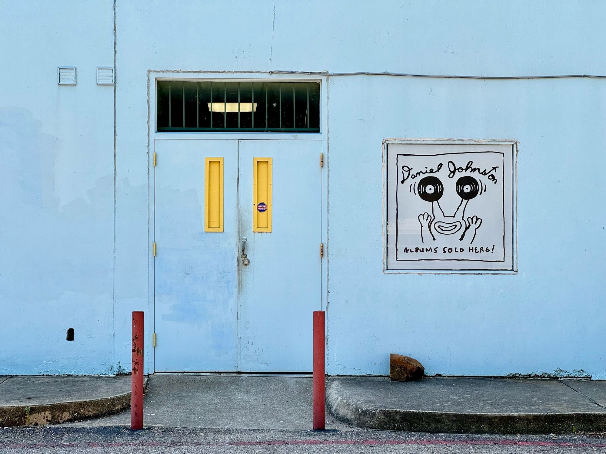 A baby blue wall in the back of the end of an ear record store in Austin Texas. Pictured next to the back door is a Daniel Johnston poster of Jeremiah the frog with records for eyes “Daniel Johnston albums sold here” it reads