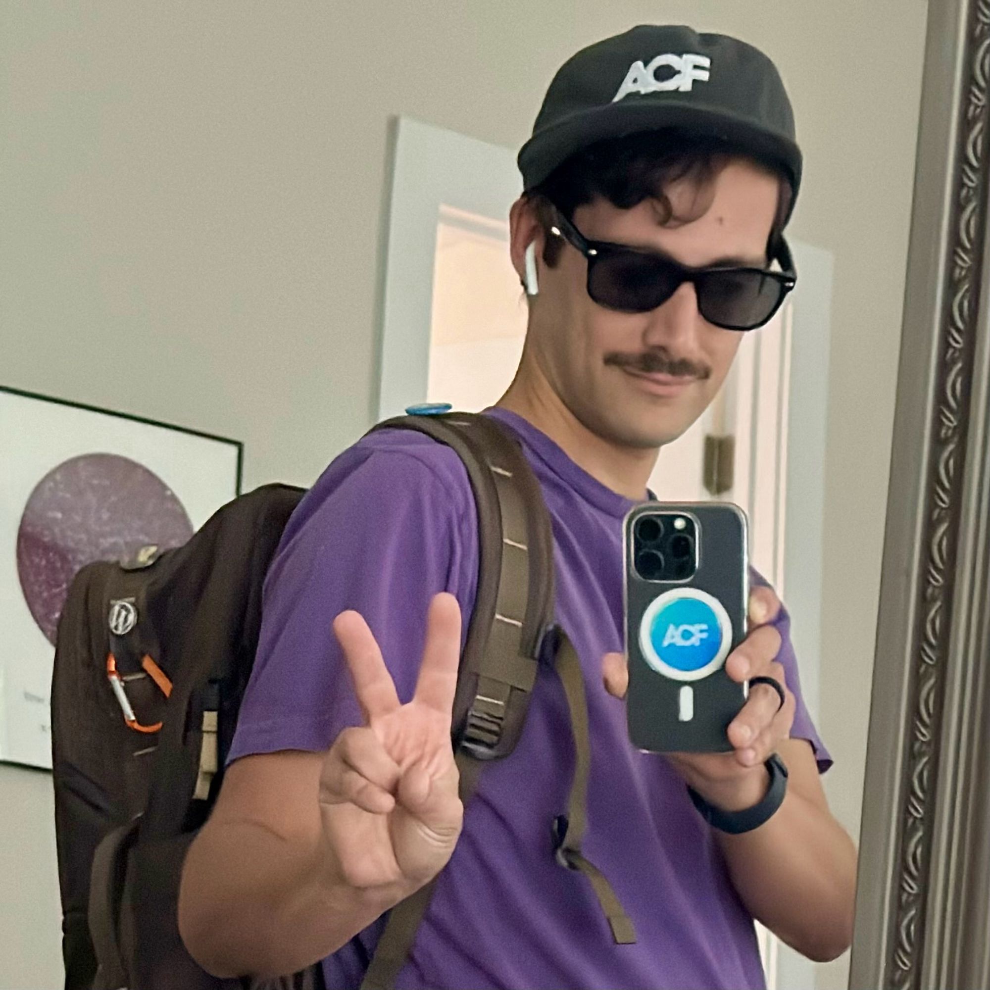 Selfie mirror shot wearing sunglasses . throwing a peace sign ✌️ and an ACF sticker center of the phone. Wearing an exclusive team ACF hat. Brown backpack with WordPress button attached. AirPods in ear because today I’m that guy. Oh and how could I forget, a mustache that I will threaten every morning with a razor.