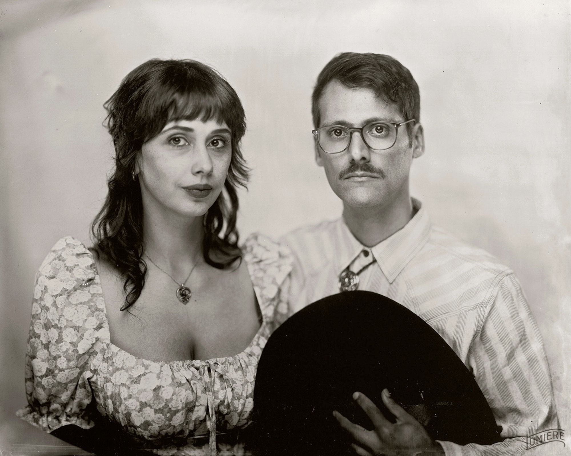 Two old timey lookin cuties. Cowboy hat over heart, mustache, wife has a floral dress and a heart and rose shaped necklace. Beautiful as ever