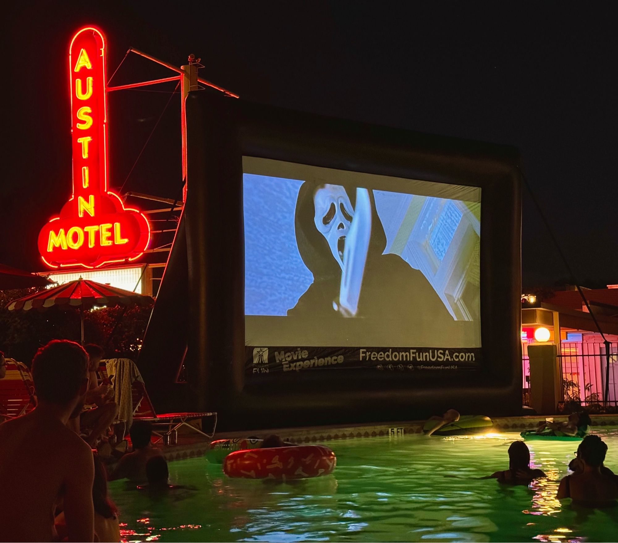 Austin Motel pool on south congress watching Scream on a big screen poolside. Pictured is a scene where ghost face has a knife out in front of his mask. Neon sign “Austin motel” lit up