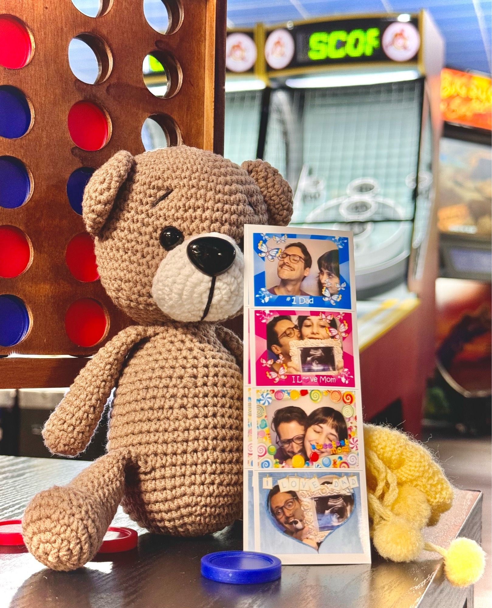 A crocheted teddy bear holding a Photo Booth strip with pictures of my wife and I holding an ultrasound with little baby us pictured! Setting is in an arcade.