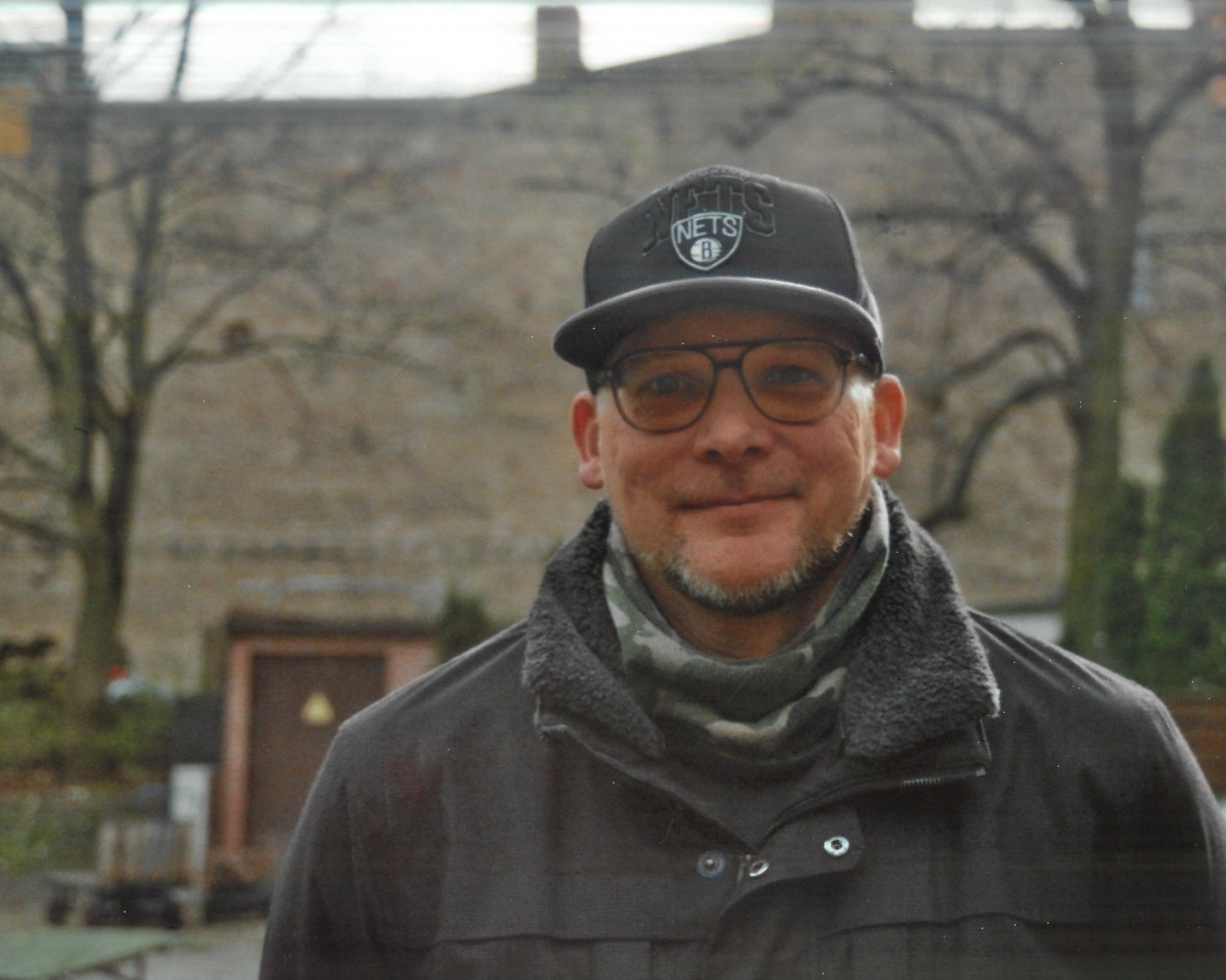 Foto zeigt mich, grinsend. Ich trage eine schwarze Cap, Brille, einen Schal und eine schwarze Jacke. Im Hintergrund eine graue Wand und zwei Bäume.