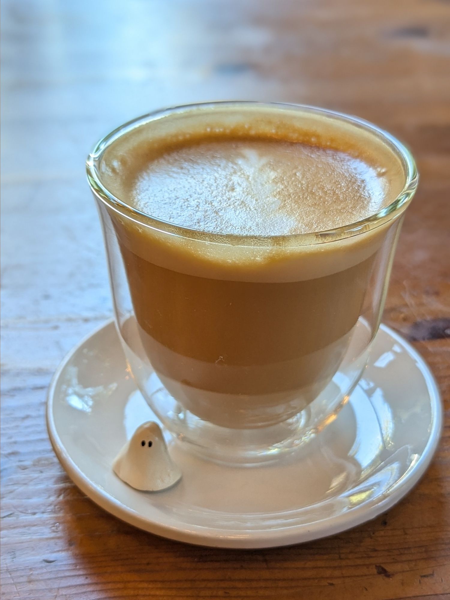 A cappuccino in a clear glass on a white plate on a wooden table. There's also a little ghost figurine on the plate.