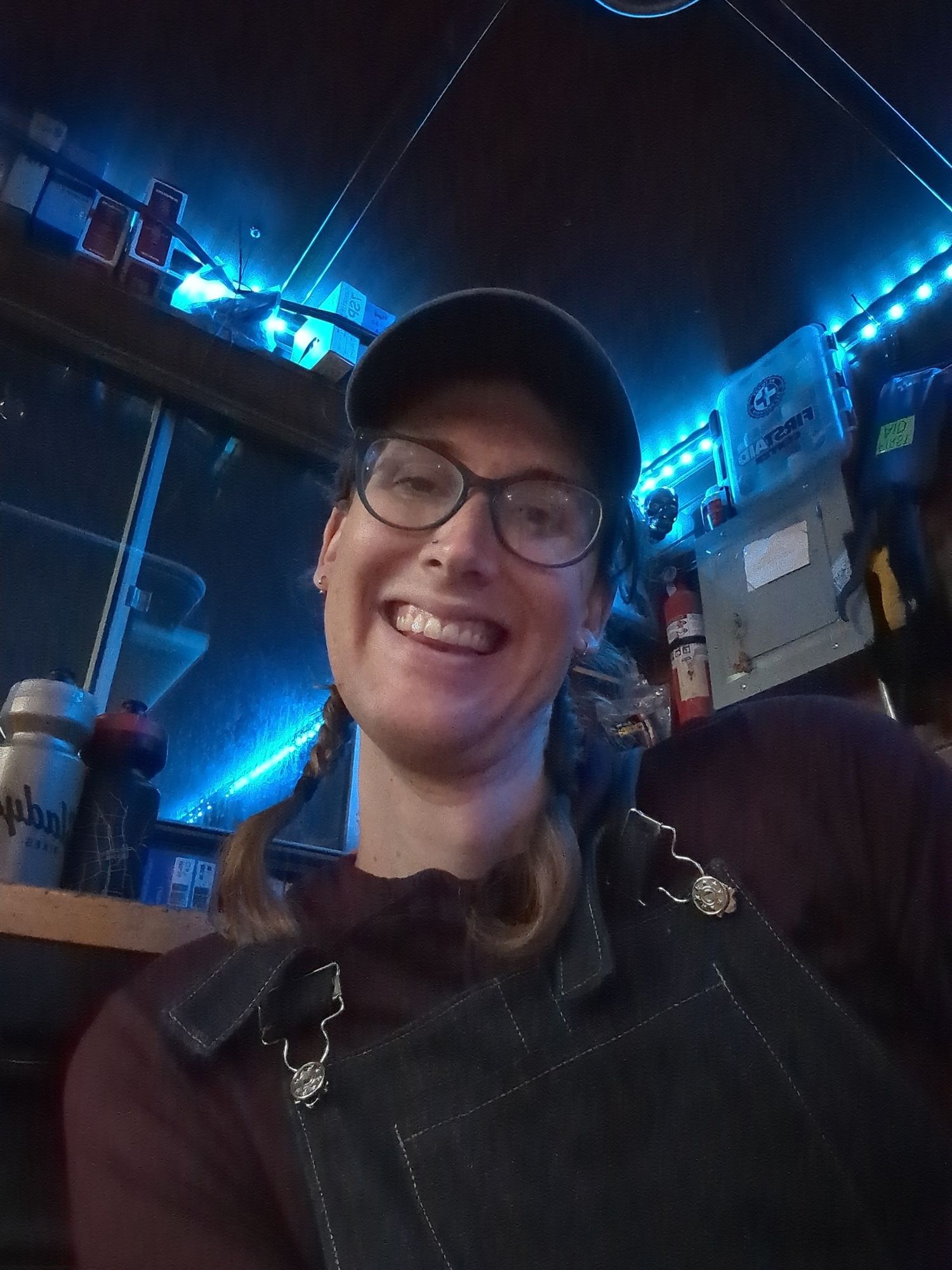 Me smiling. With braided pigtails, a blue cap, glasses, a purple shirt, and overalls. Sitting in a dark bike shop with mood lighting.