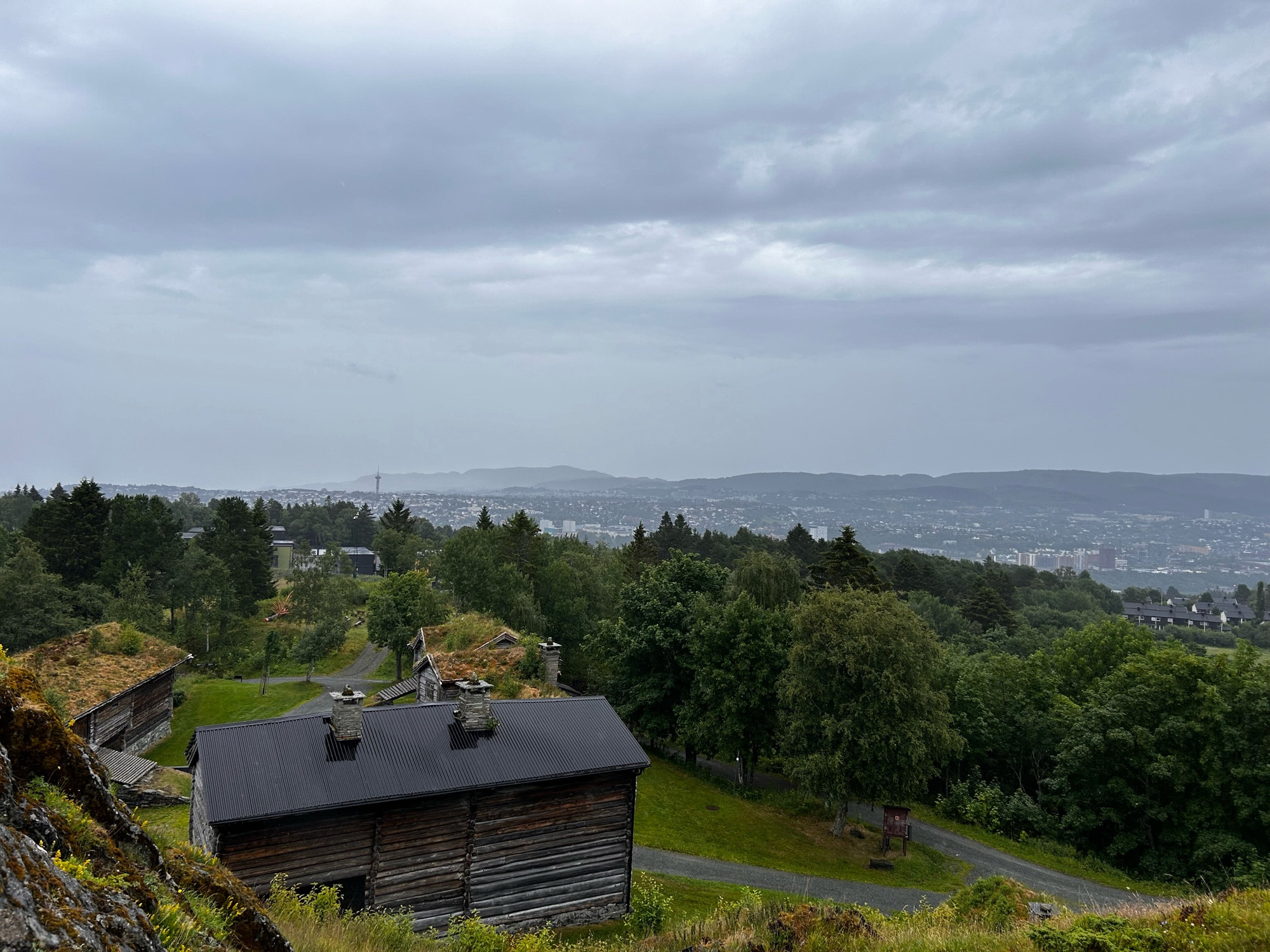 Bilde tatt fra borgplatået på Sverresborg. I forgrunnen står noen gamle laftahus, noen med torvtak. En rekke med løvtrær i midten og Trondheim i bakgrunnen. Tyholt tårnet vises så vidt til venstre.