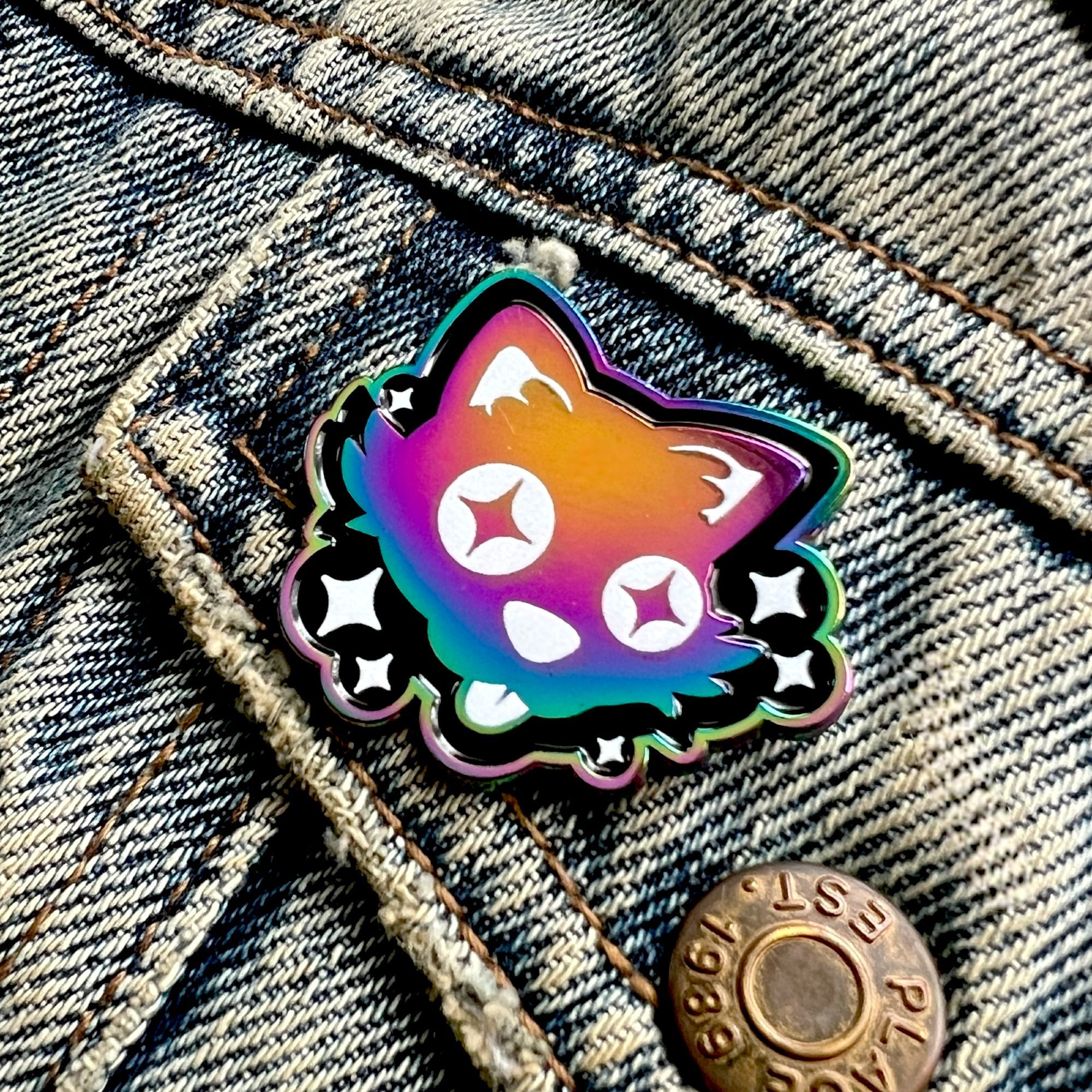 A close up of the anodised rainbow enamel pin of Cosmic Cat, a starry-eyed kitty with his tongue sticking out