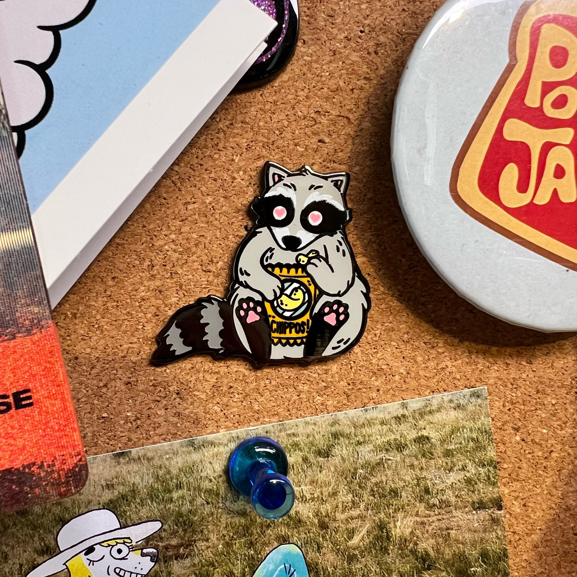 A cork board, the central focus of which is an enamel pin of a fat raccoon with heart eyes eating Chips (labelled chippos!)