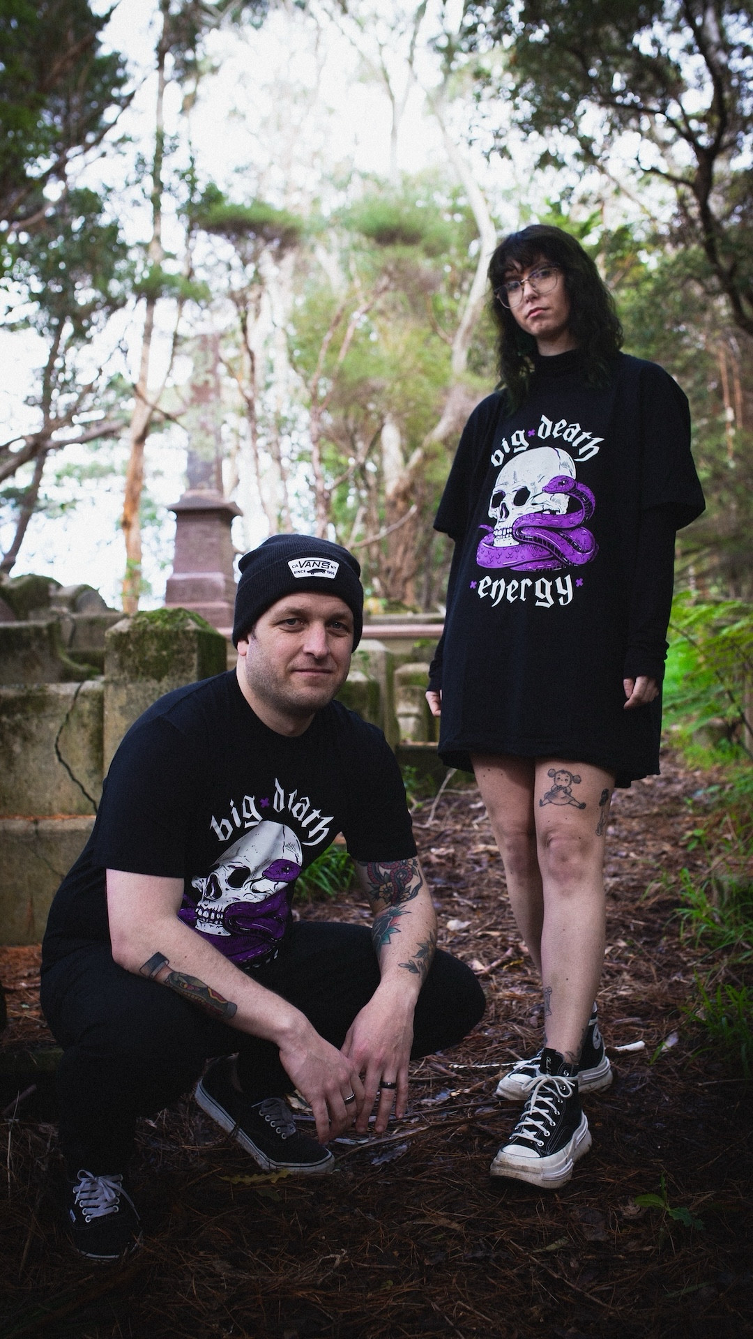 Two models wearing Pepper Raccoon tees in a graveyard. The femme presenting one is wearing a long Big Death Energy tee, platform converse, and glasses. She has tattoos and short brown wavy hair. The masc presenting one has a beanie and is crouching, also in a Big Death Energy Tee. The tee has a skull and a purple snake on it.