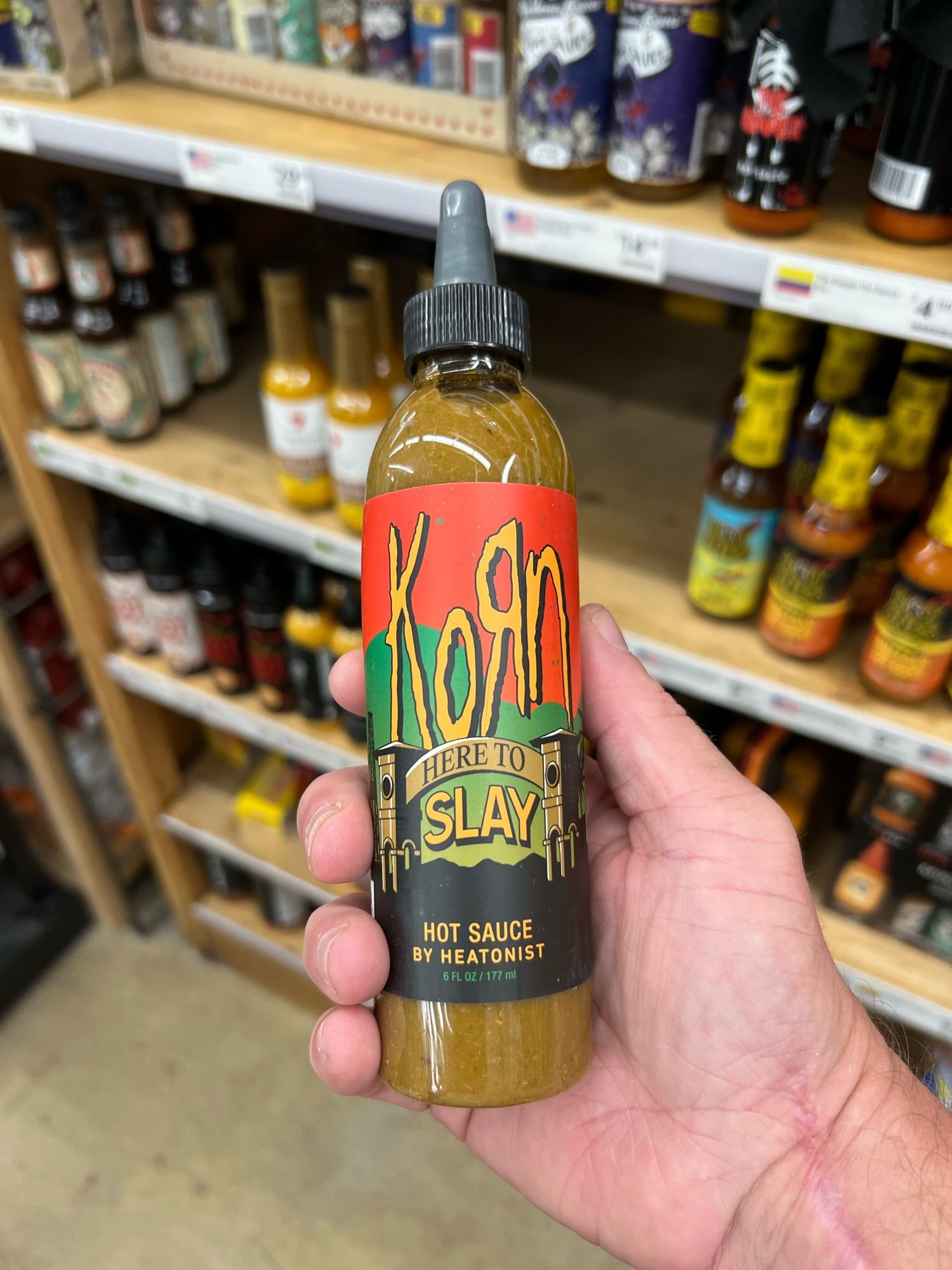 A hand is holding a bottle of hot sauce in a store. The label on the bottle reads "Korn Here to Slay" in large, stylized letters, with "Hot Sauce by Heatonist" below. The label has a red and black gradient background with some green accents, and two architectural columns are depicted on each side of the word "Slay." The bottle contains a light green sauce and has a black screw-on cap with a pointed applicator tip. Shelves in the background display various other hot sauce bottles.