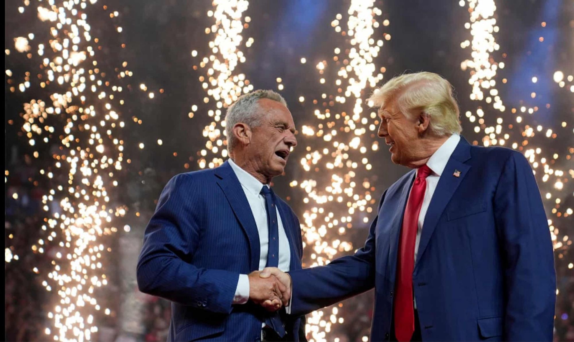 The image shows Robert F. Kennedy Jr. and Donald Trump shaking hands while facing each other. RFK Jr., on the left, has gray hair and is wearing a blue pinstripe suit with a white shirt and dark tie. Donald Trump, on the right, has blond hair and is dressed in a dark blue suit with a white shirt and red tie. They are standing against a backdrop of fireworks, which fill the background with bright, scattered bursts of light.