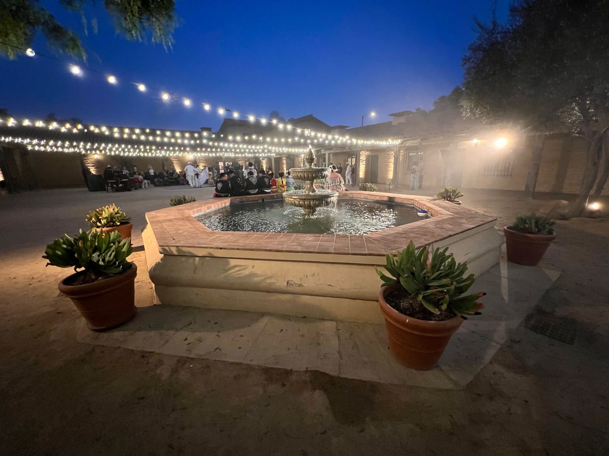 The image depicts an evening outdoor scene with a square-shaped fountain at the center. The fountain is surrounded by a tiled edge and four large potted plants, each filled with green leafy plants. Overhead, strings of lights are hung in a crisscross pattern, illuminating the courtyard with a soft, warm glow. In the background, people are seated at tables, gathered in a relaxed, social setting. The sky is a deep blue, indicating dusk, and a tree with branches hanging into the frame is partially visible in the upper left corner. The overall ambiance is warm and inviting.